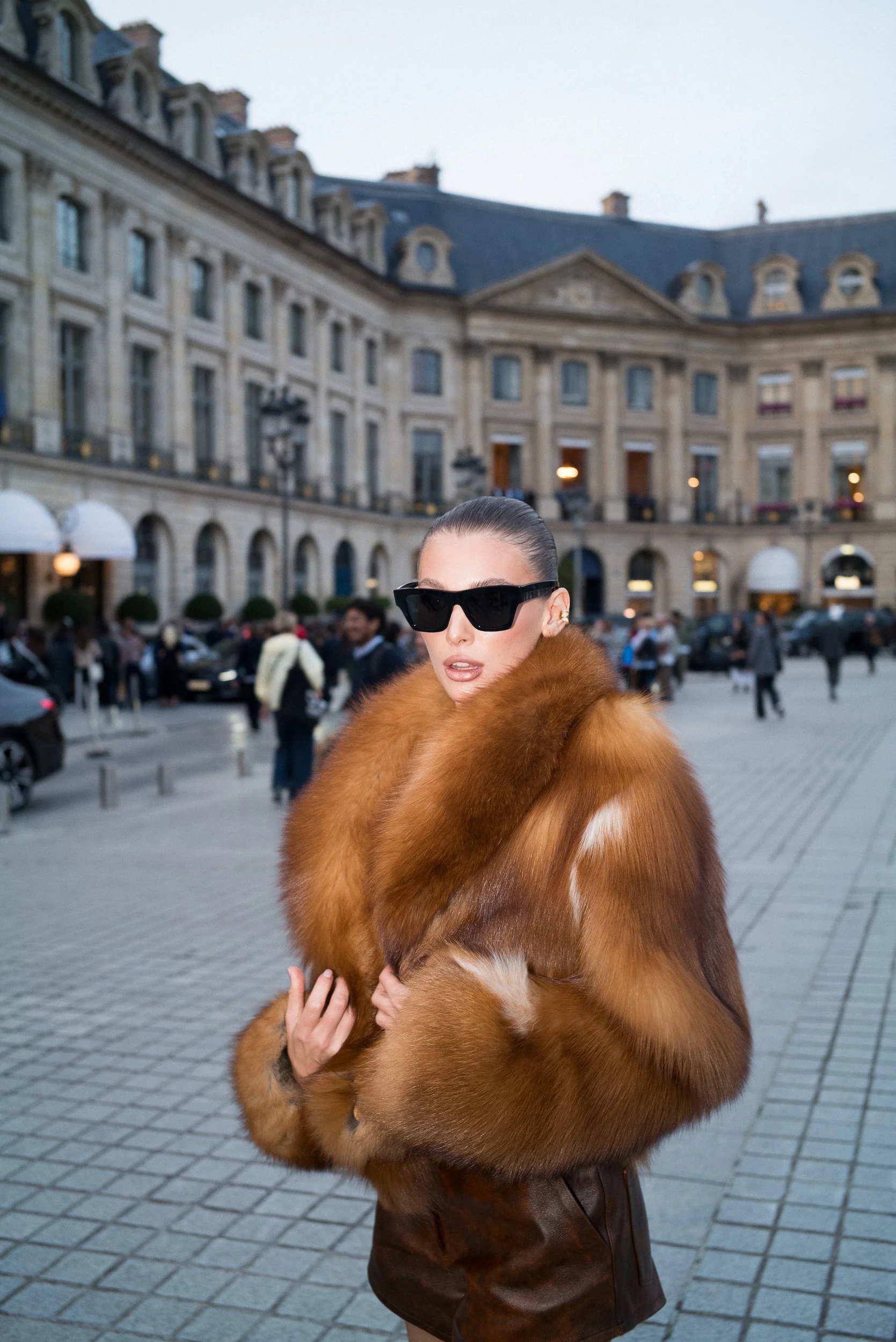 At Paris Fashion Week, A woman in a large fur coat and black sunglasses standing outdoors in a city square with historic buildings and many people in the background.
