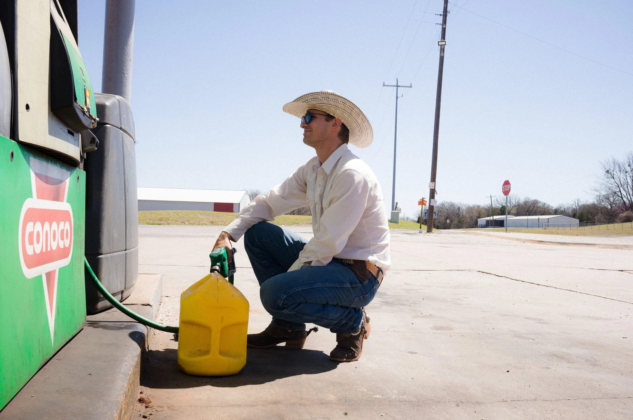 Man kneeling outside at a gas pump with a yellow container nearby, wearing a large straw hat, sunglasses, a beige shirt, and jeans, filling a container with fuel on a sunny day with utility poles and a stop sign in the background.
