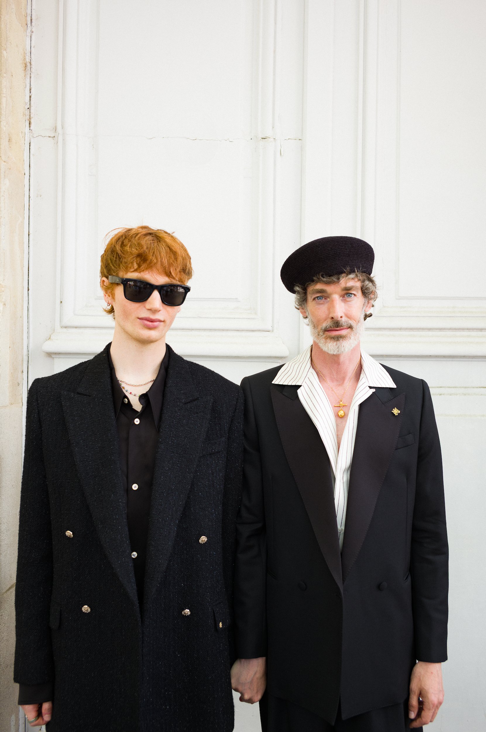 Two individuals standing in front of a white wall. The person on the left has short red hair, wearing sunglasses, a black blazer, and a black shirt. The person on the right has curly gray hair, wearing a black beret, a black blazer with a pin, a whit