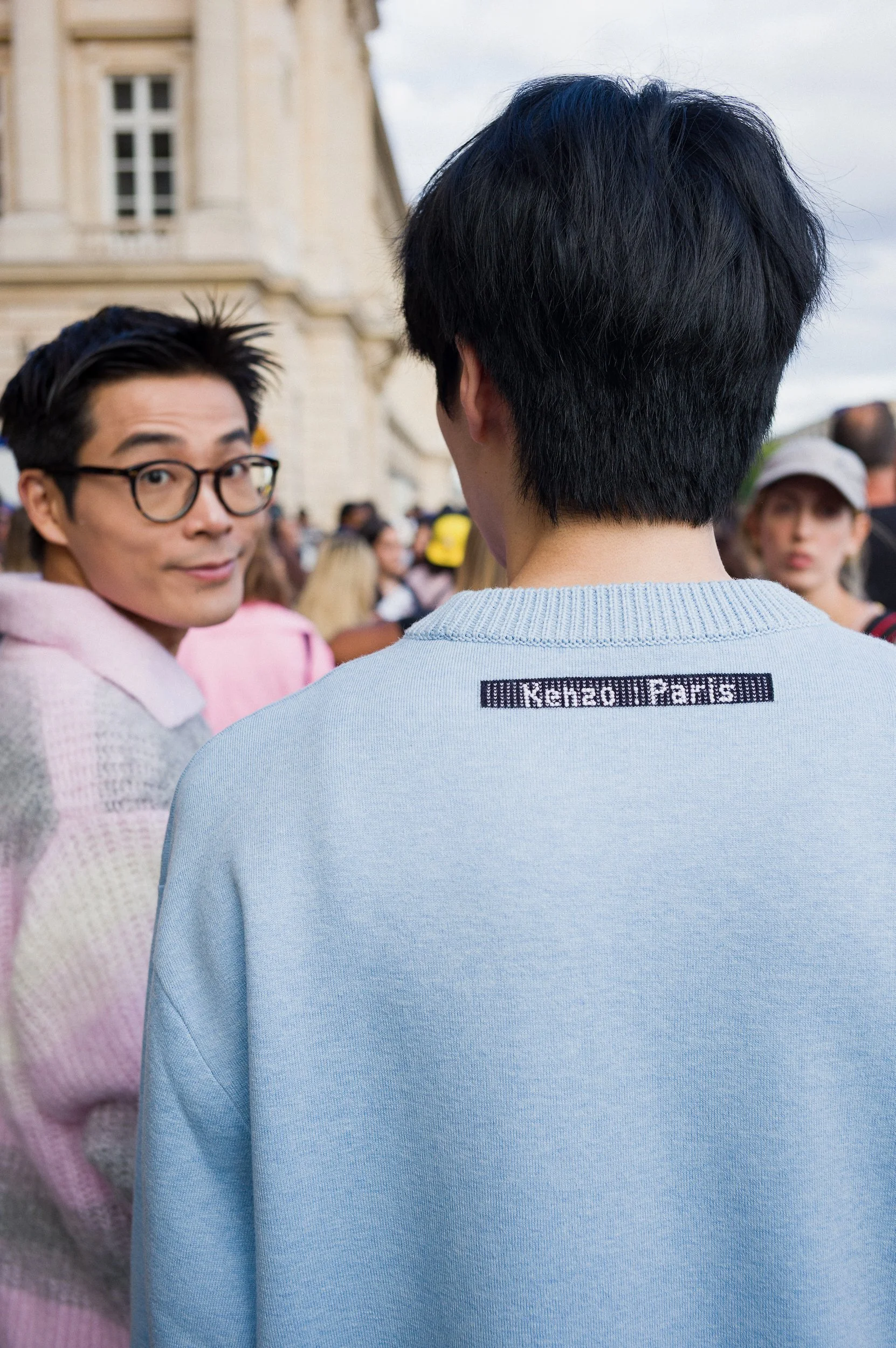 A person with black hair wearing a light blue sweater with black text on the back, standing outdoors during daytime. In the background, a crowd of people and a historic building are visible, with one person wearing glasses looking back on the left si