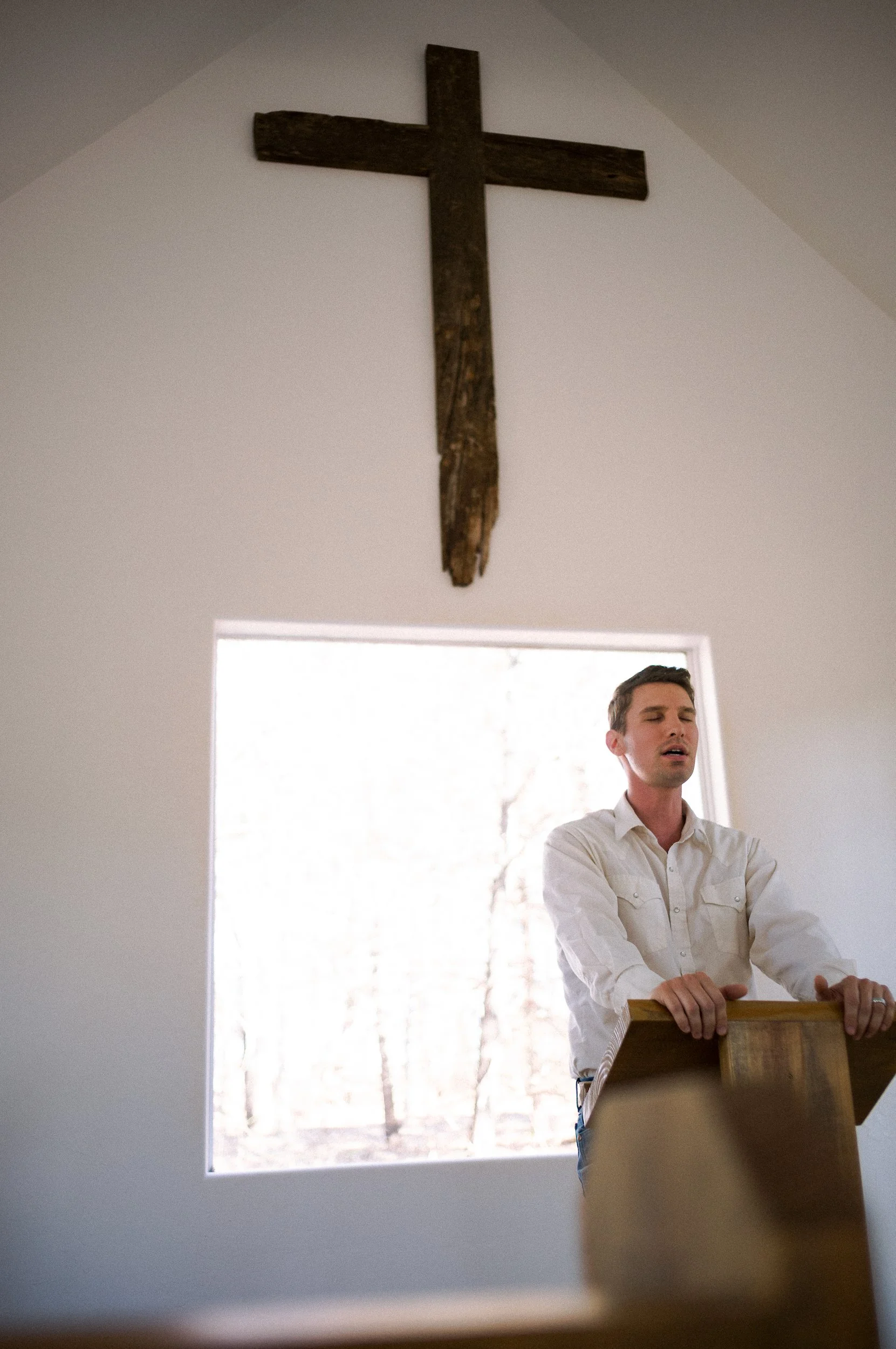 A man standing at the front of a chapel, with his eyes closed and hands resting on the podium, inside a room with white walls and a large window showing trees outside. A wooden cross hangs on the wall above him.