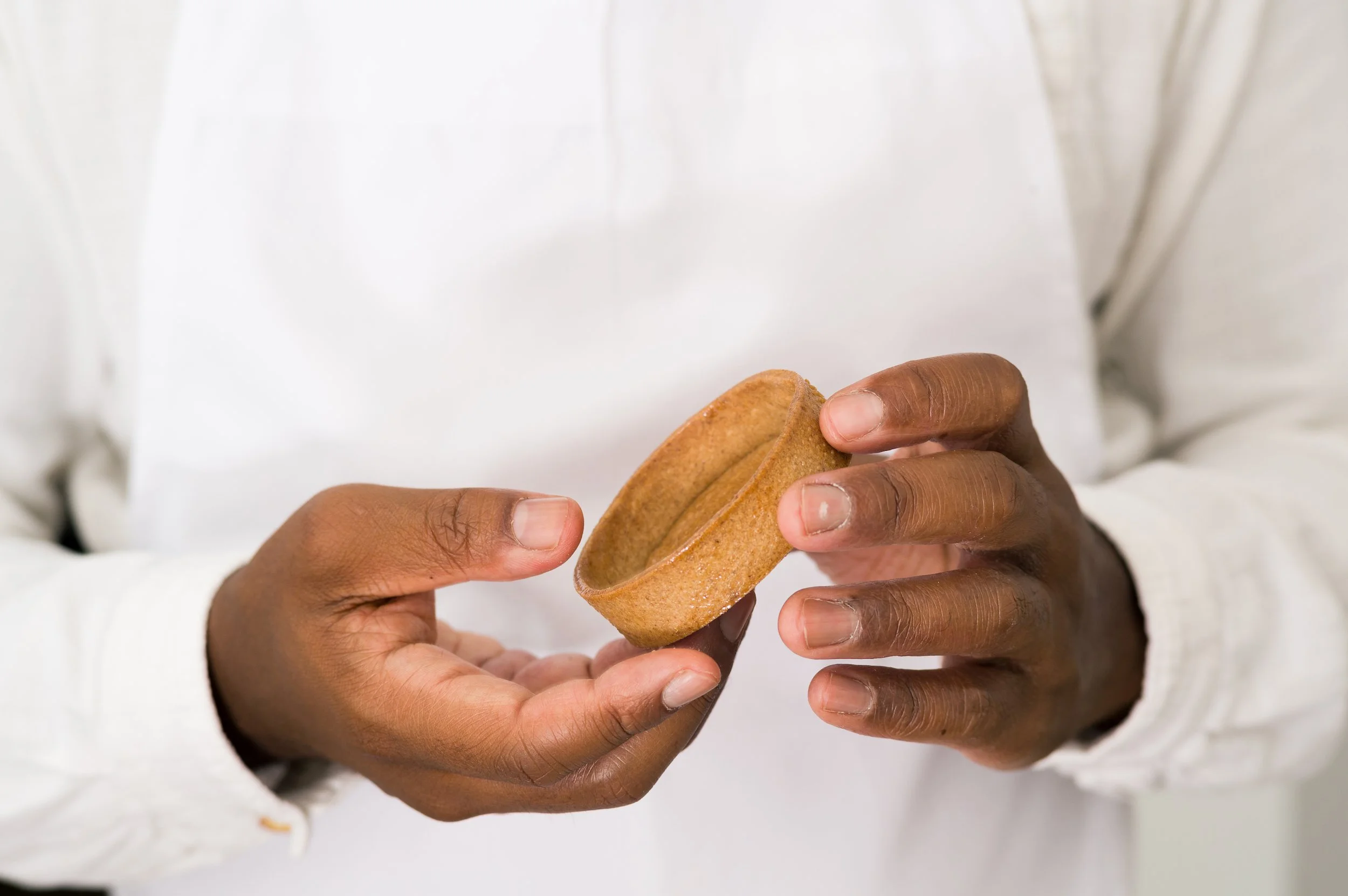 A chef with dark skin holding a small, oval-shaped cookie or biscuit with both hands. The person is wearing a white garment, and the background is plain white.