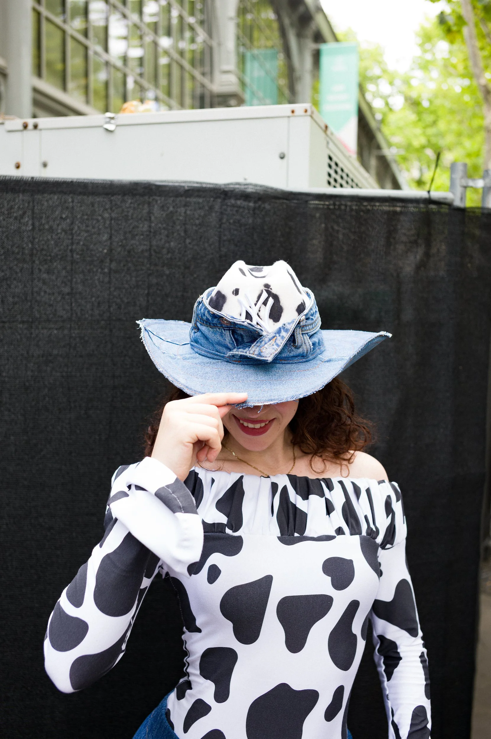 At Paris Fashion Week, A woman wearing a cow-patterned hat and matching long-sleeve top, standing outdoors in front of a black background and covering part of her face with her hand.