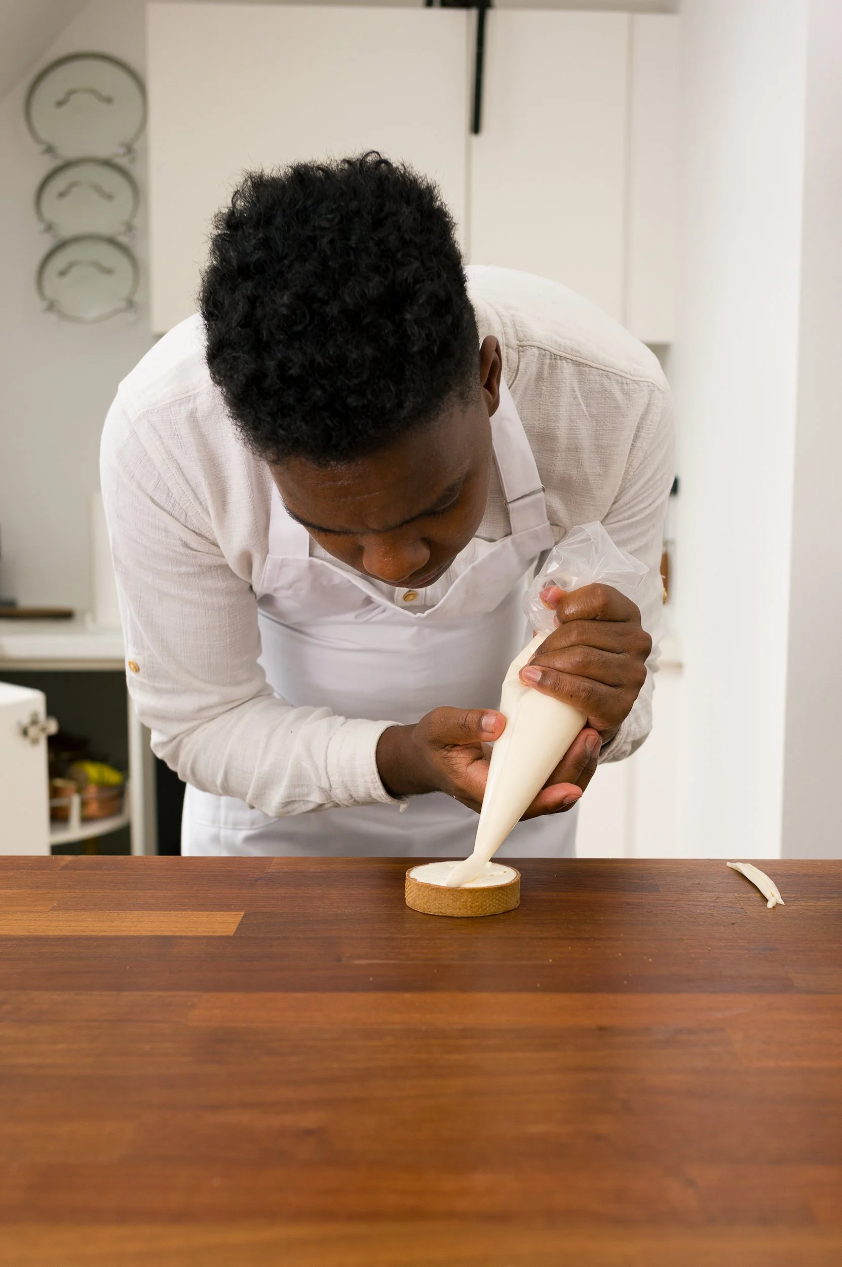 A chef is pouring cream onto a cake in a kitchen, wearing a white shirt and apron, with a focused expression.