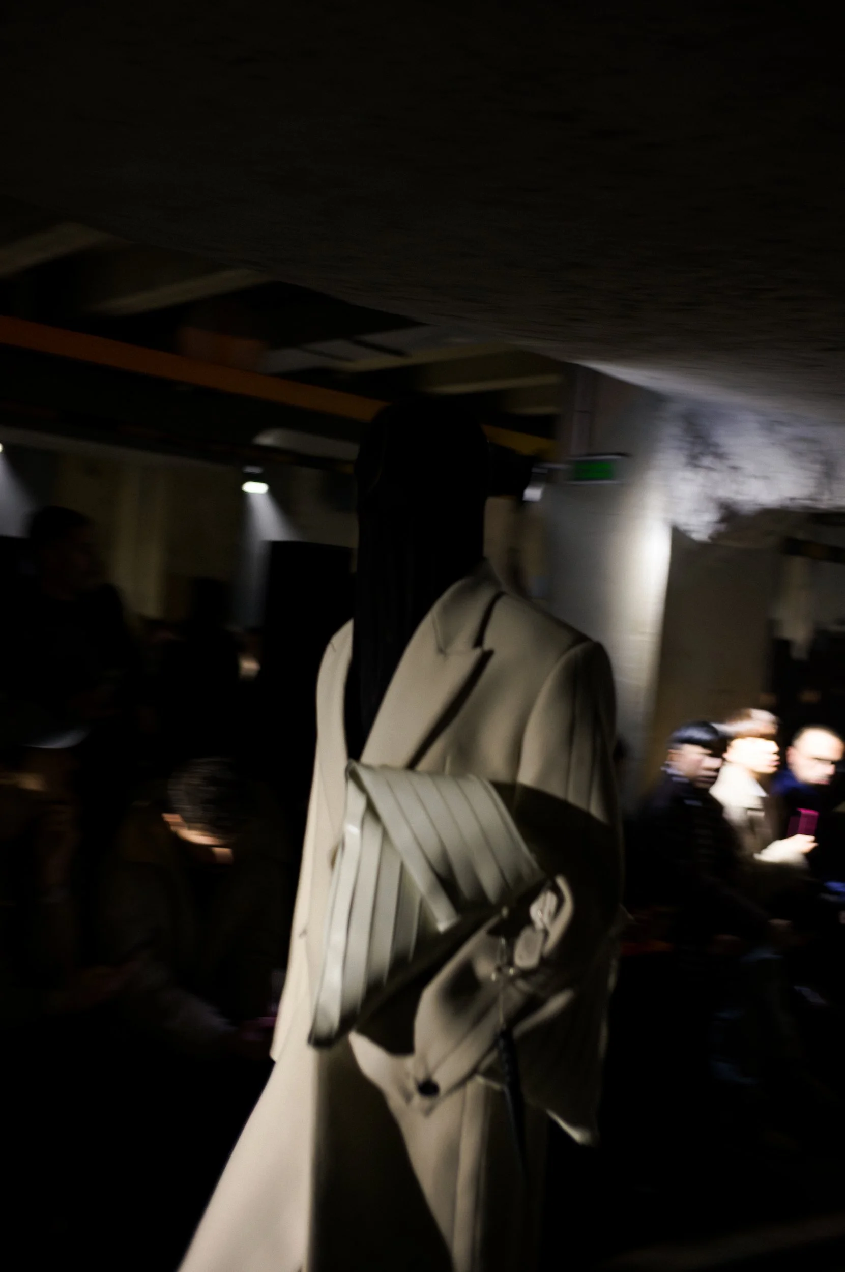 At Paris Fashion Week, A person with long black hair carrying a large white and black strap backpack walking through a dimly lit indoor space with other people around at the Louis Gabriel Nouchi Runway.