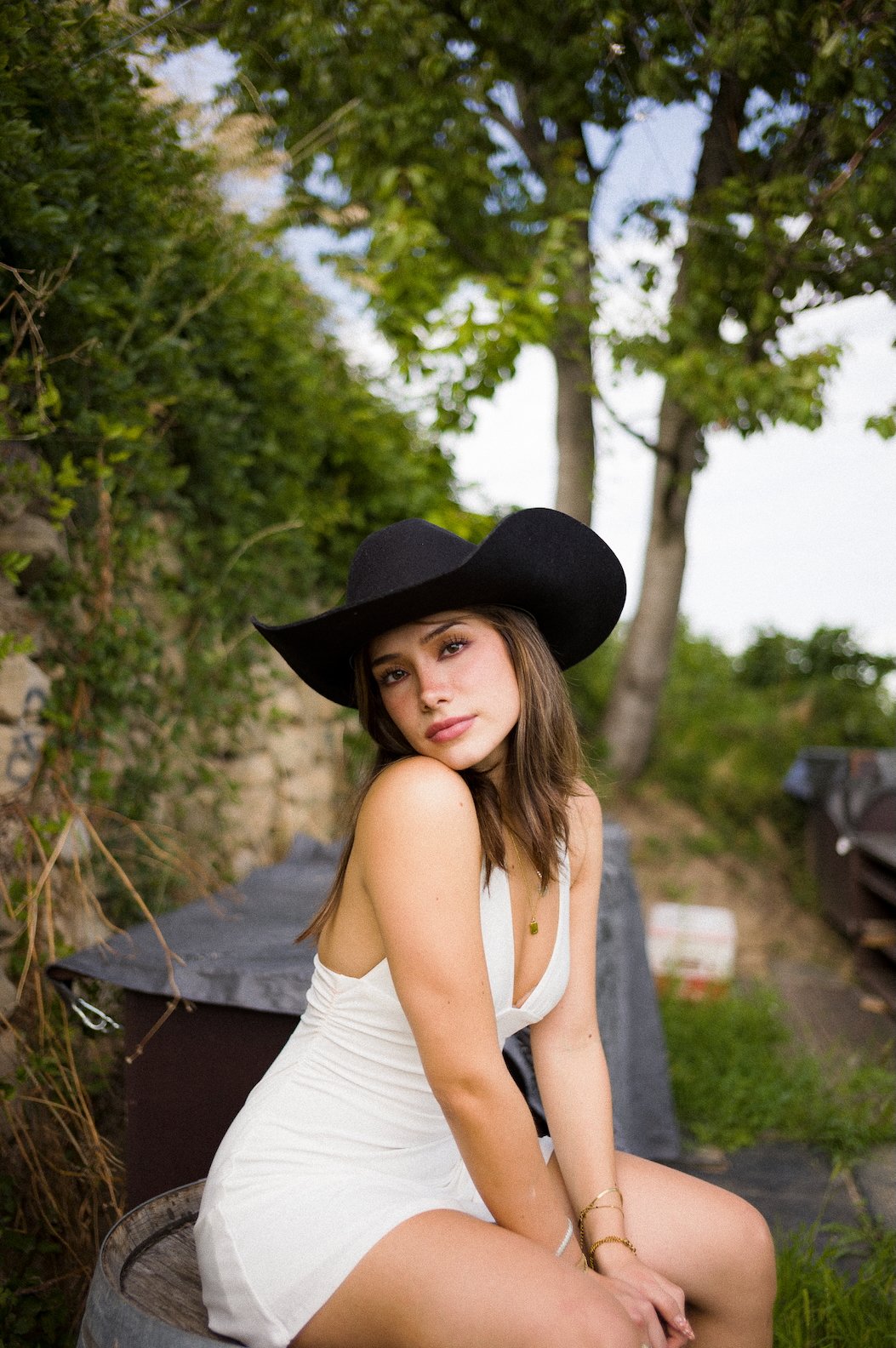A young woman wearing a black wide-brimmed cowboy hat and a white sleeveless dress, sitting outdoors with trees and greenery in the background.