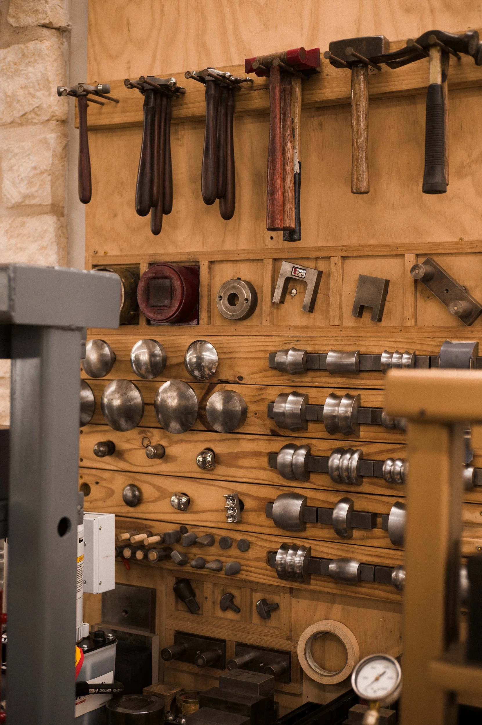 Wall-mounted silver working tools including various hammers, mallets, and metalworking implements on a wooden board in a workshop.