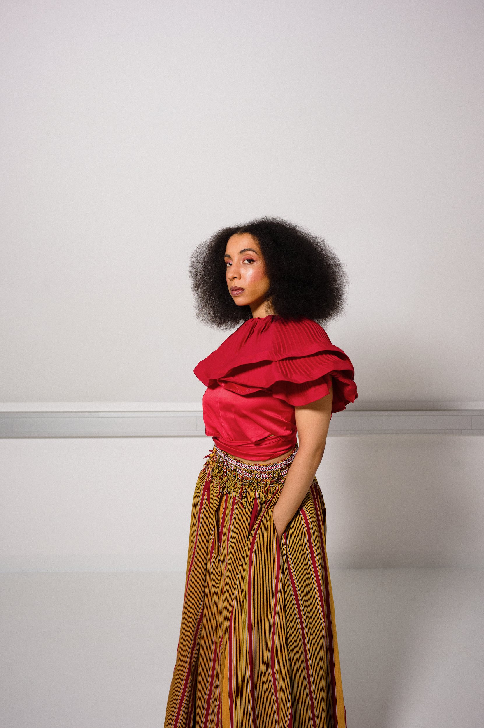 Woman with dark, curly hair wearing a red ruffled top and colorful striped pants standing against a plain white background.