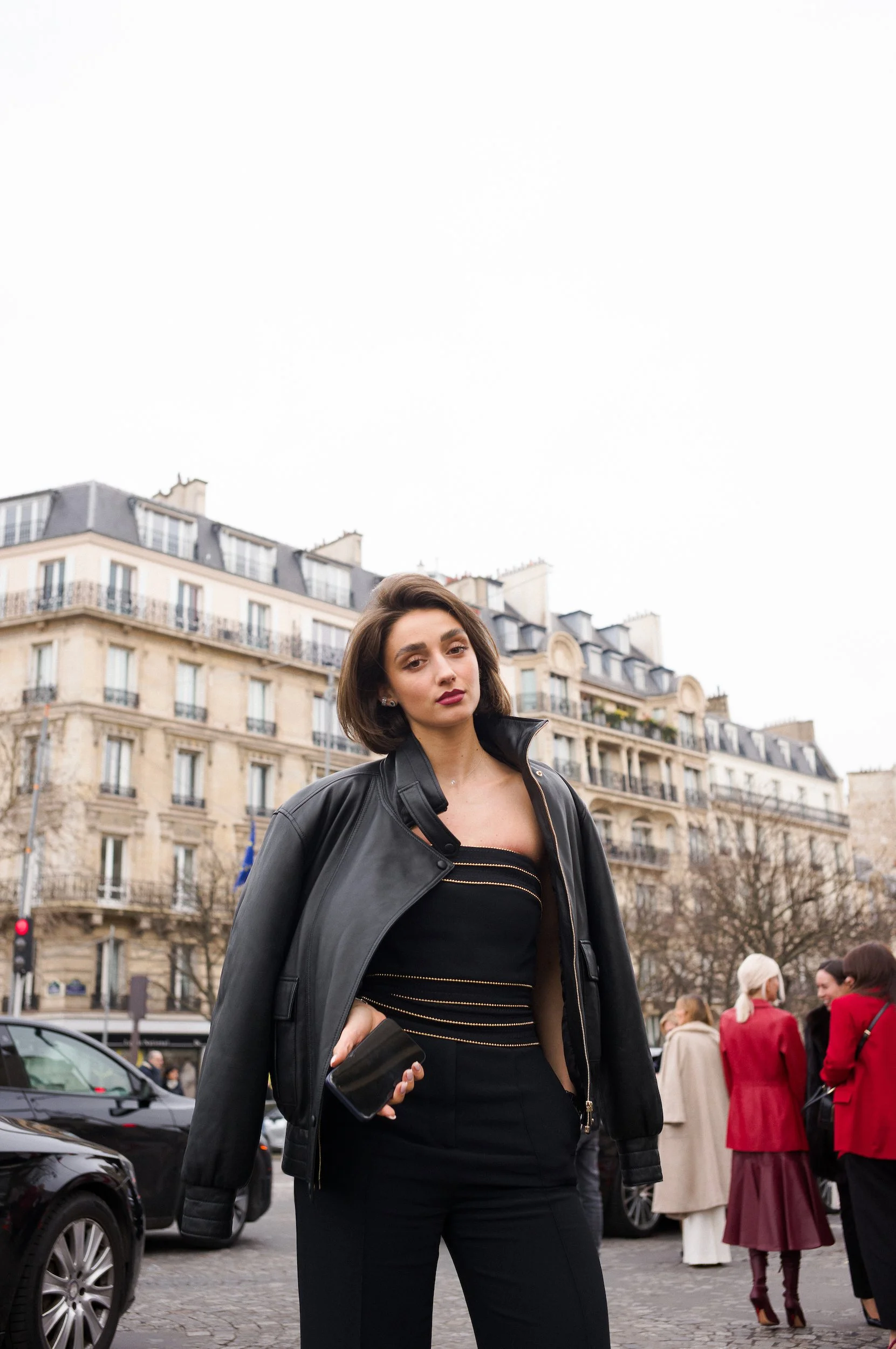 A woman with dark hair and red lipstick standing on a city street, wearing a black leather jacket and black pants, holding a black clutch, with classic Parisian buildings and other pedestrians in the background.