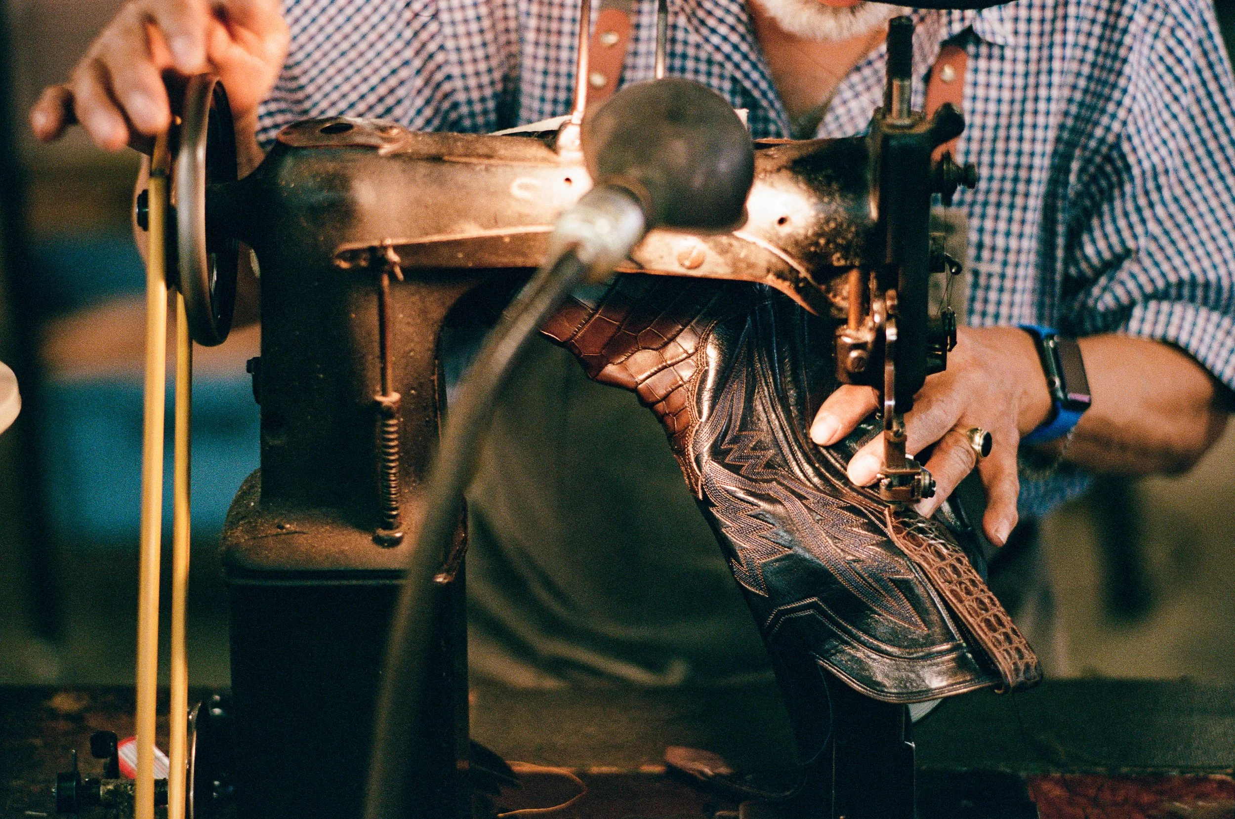A person sewing leather cowboy boots using a sewing machine. The person is holding the shoe with one hand while operating the machine with the other. The person's face is not visible, and they are wearing a checkered shirt and a wristwatch.