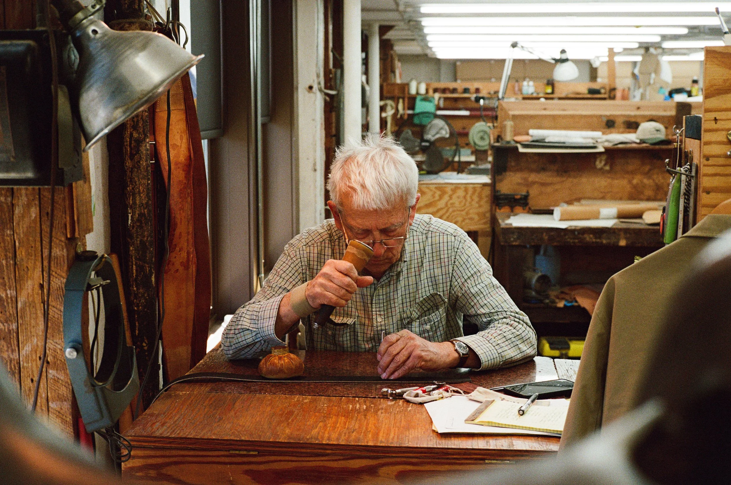 In ML Leddys, an elderly man with white hair and glasses is sitting at a wooden table in a workshop, carefully working on a leather belt using a magnifying glass. The workshop is filled with various tools and wooden shelves in the background.