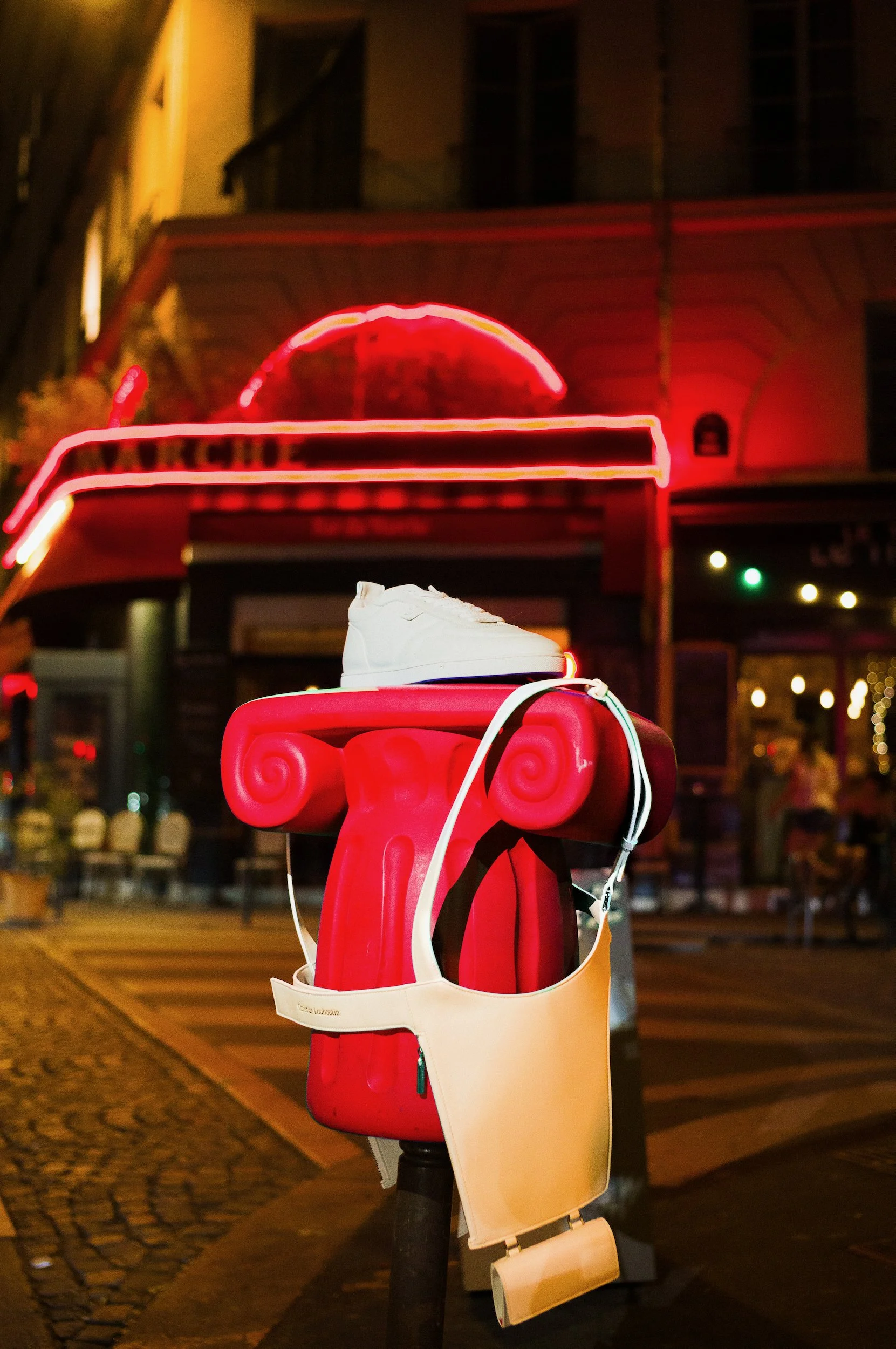 A white sneaker placed on top of a red and beige decorative object, with a city street background illuminated by neon lights at night.