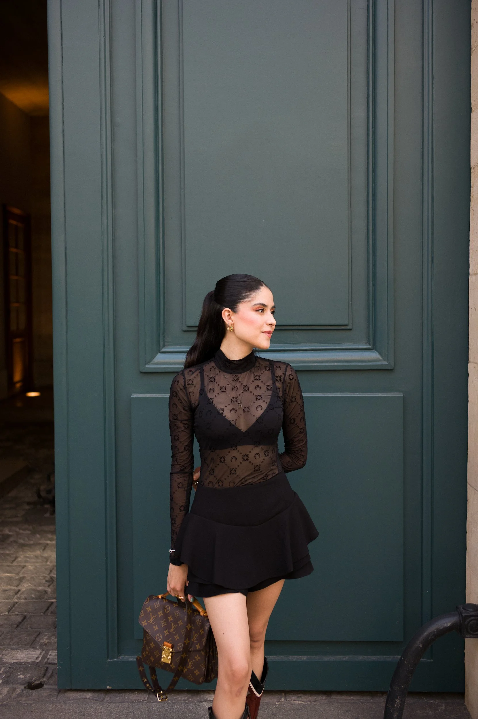 A woman with dark hair, wearing a black lace long-sleeve top and a black skirt, is standing in front of a large dark green paneled door. She is holding a Louis Vuitton backpack in one hand and wearing black boots. The woman is looking to her left, wi