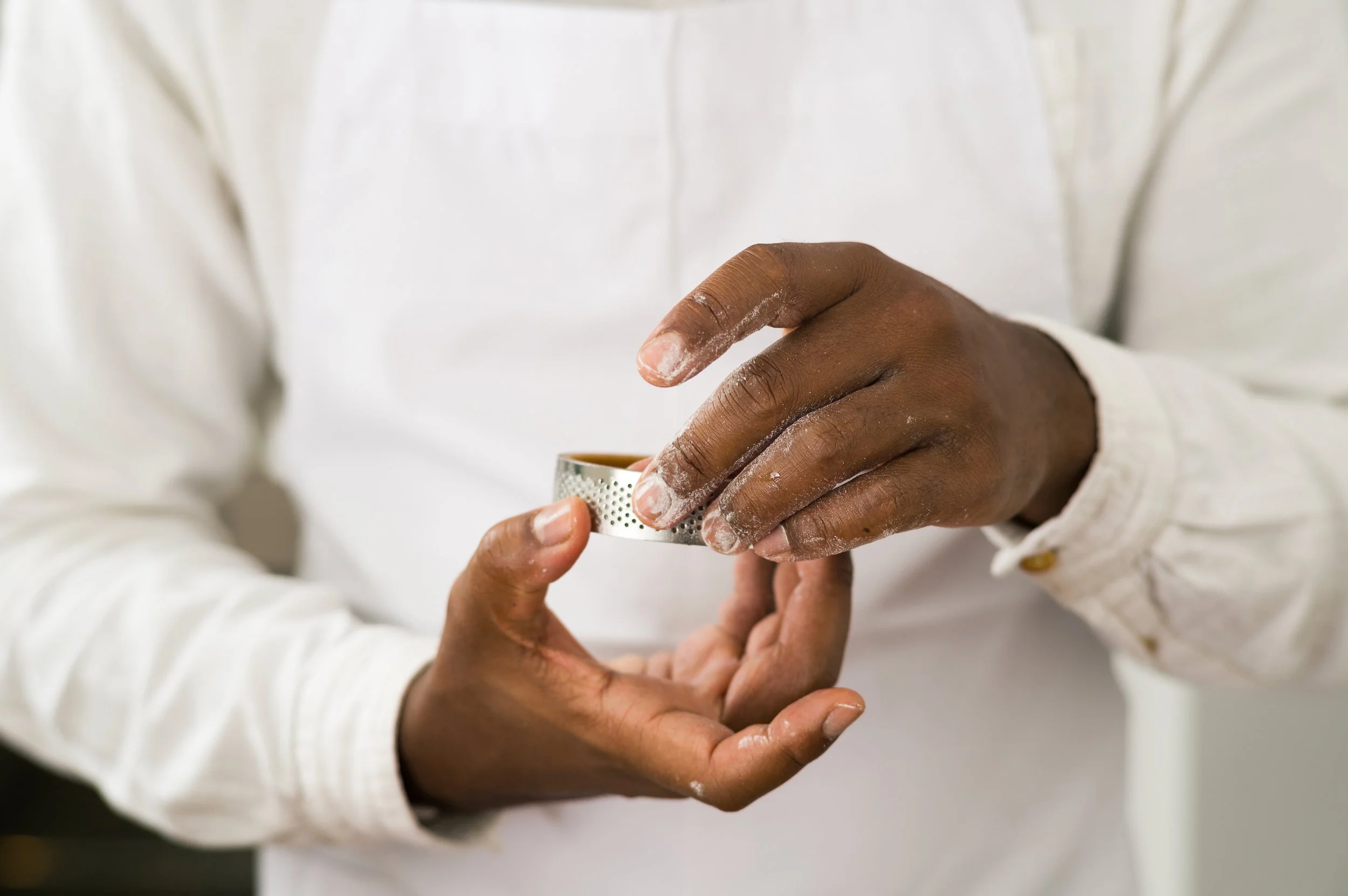 A chef's hands holding a small metal grater, with one hand on top and the other supporting below. The person is wearing a white long-sleeve shirt, and some flour or powdered substance is visible on their fingers.