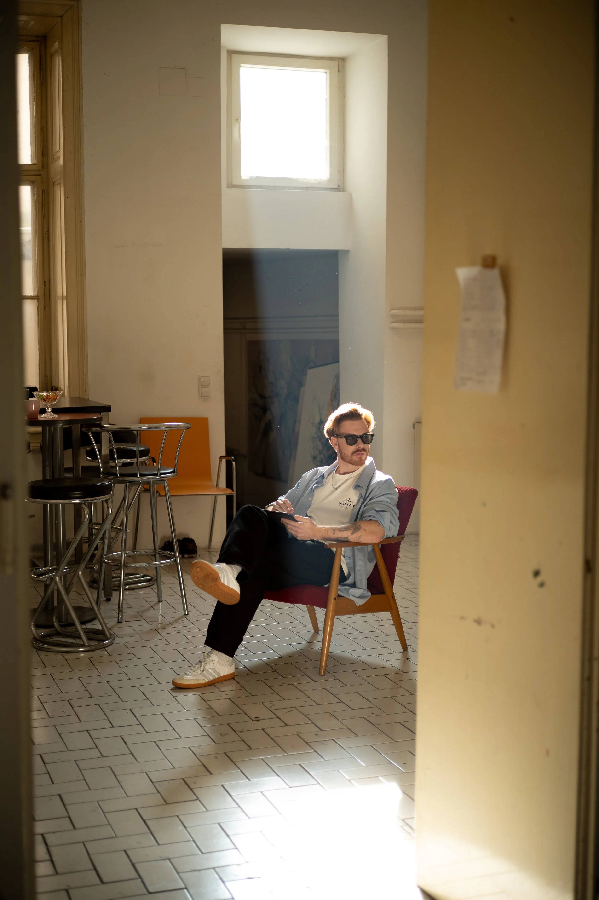 A man with glasses, sitting in a red chair, wearing a light blue shirt, white sneakers, and black pants, holding a device, in a room with yellow and white walls, tiled floor, and barstools.