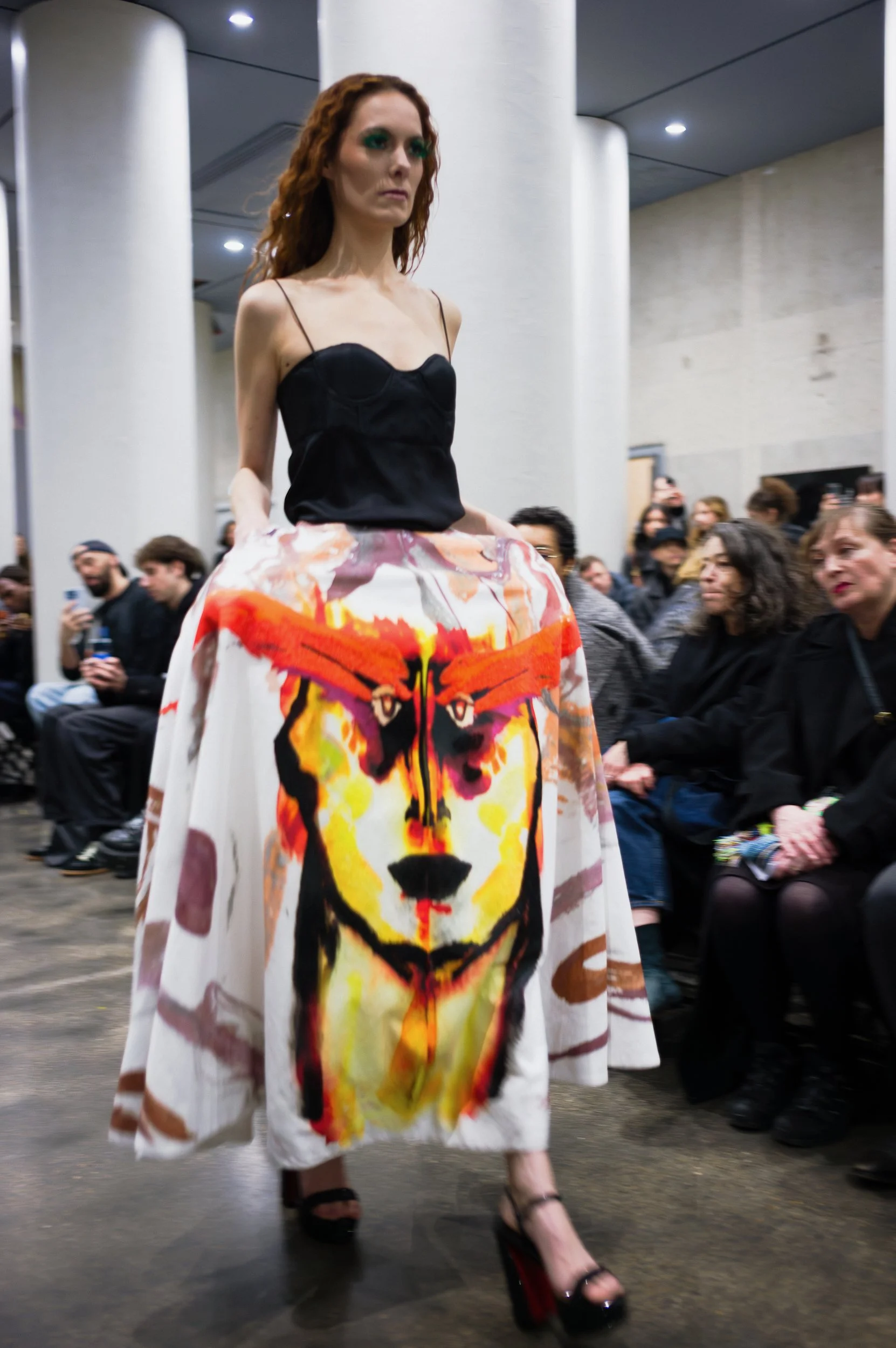 At Paris Fashion Week,  A woman walking for Valette Studio fashion show, wearing a long white skirt with a vivid tiger face print and a black camisole top. The audience is seated on either side of her in an indoor venue with white columns.