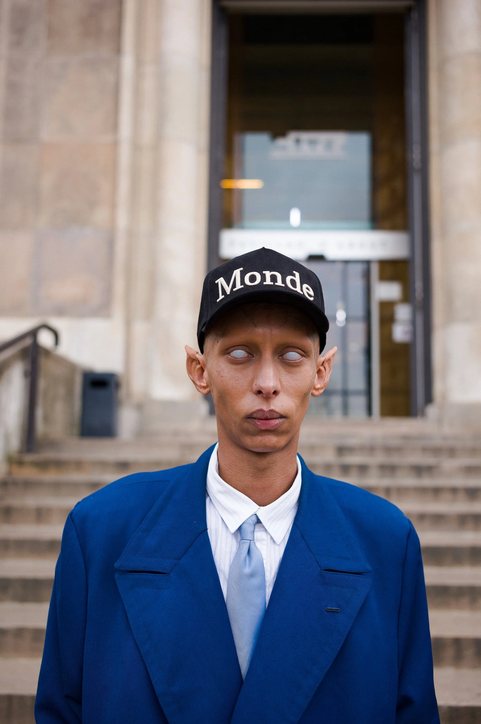 At Paris Fashion Week, A person with exaggerated facial features wearing a black cap with white lettering, a blue suit, and a white shirt, standing in front of a building with stairs and an entrance doorway.