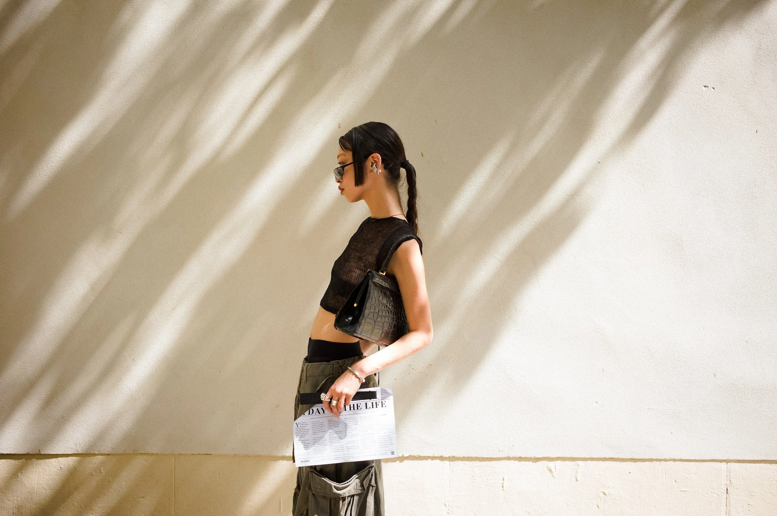 A young woman with dark hair tied in a braid wearing sunglasses and earrings, dressed in a black sleeveless top and cargo pants, standing sideways against a beige wall with shadow patterns. She is holding a newspaper titled 'Day of the Life' and a bl