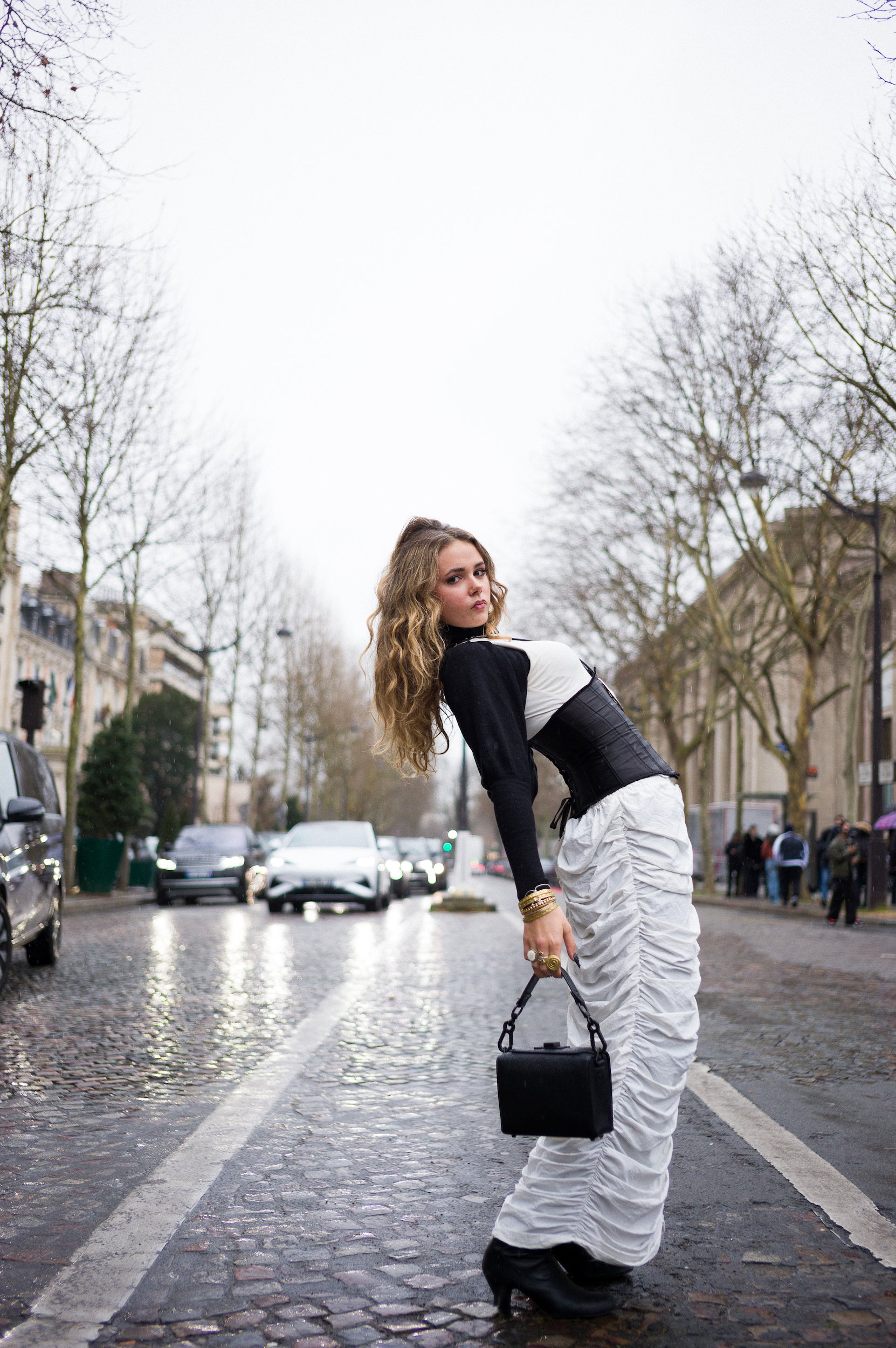 A woman with wavy blonde hair, wearing a black and white top, white ruched pants, and black high-heeled boots, standing on a cobblestone street in an urban area with leafless trees and cars in the background, holding a black handbag and looking at th