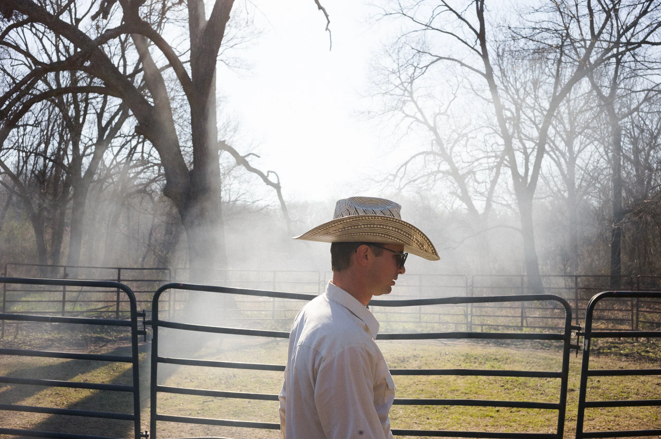 A man wearing a cowboy hat and glasses, with a white shirt, standing outdoors during foggy weather, surrounded by leafless trees and metal fencing.