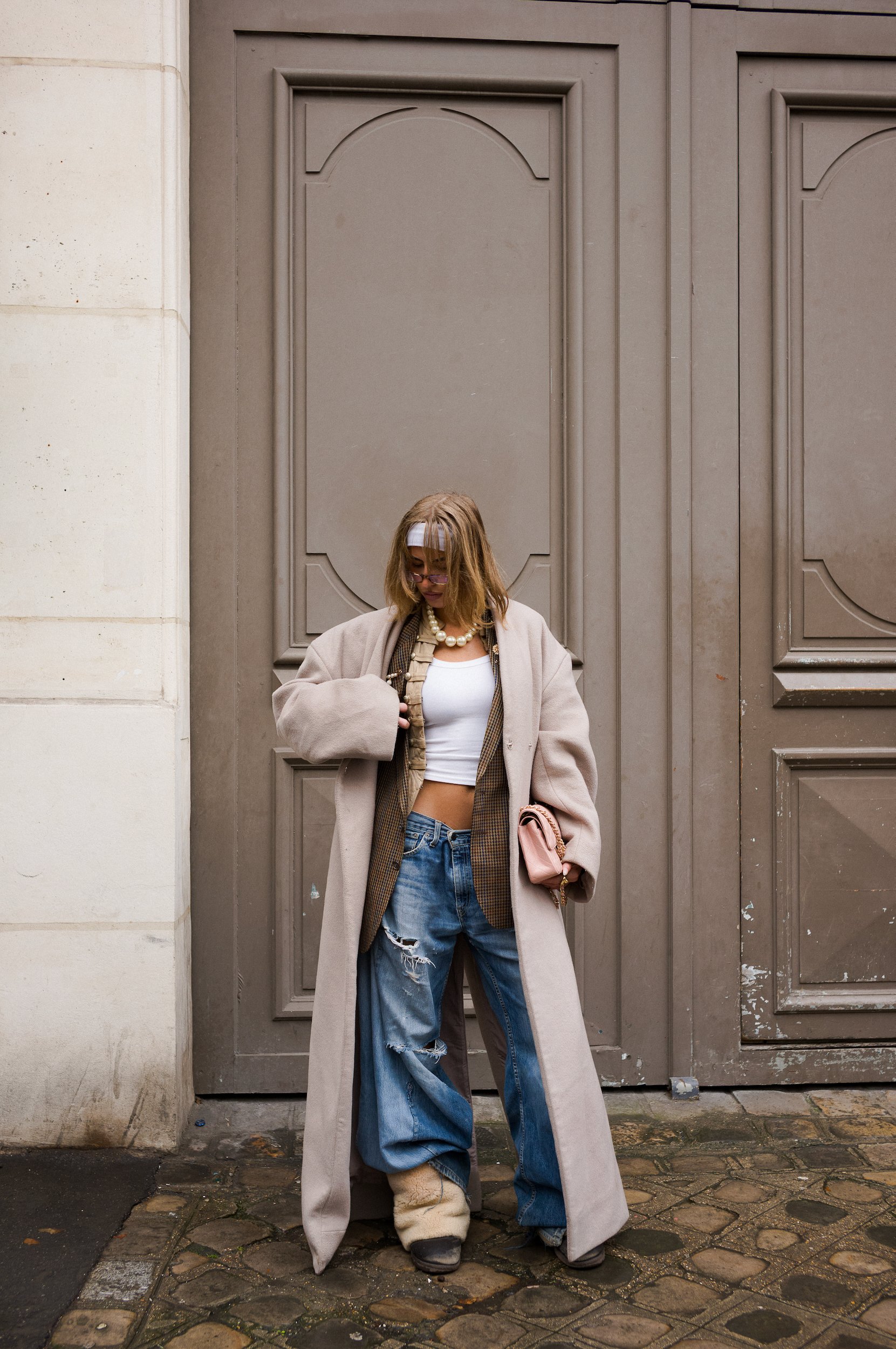 A person stands in front of a large grey door with decorative panels. They are wearing a beige coat, ripped blue jeans, a white crop top, and fluffy boots. They have shoulder-length hair, glasses, and are holding a pink bag.