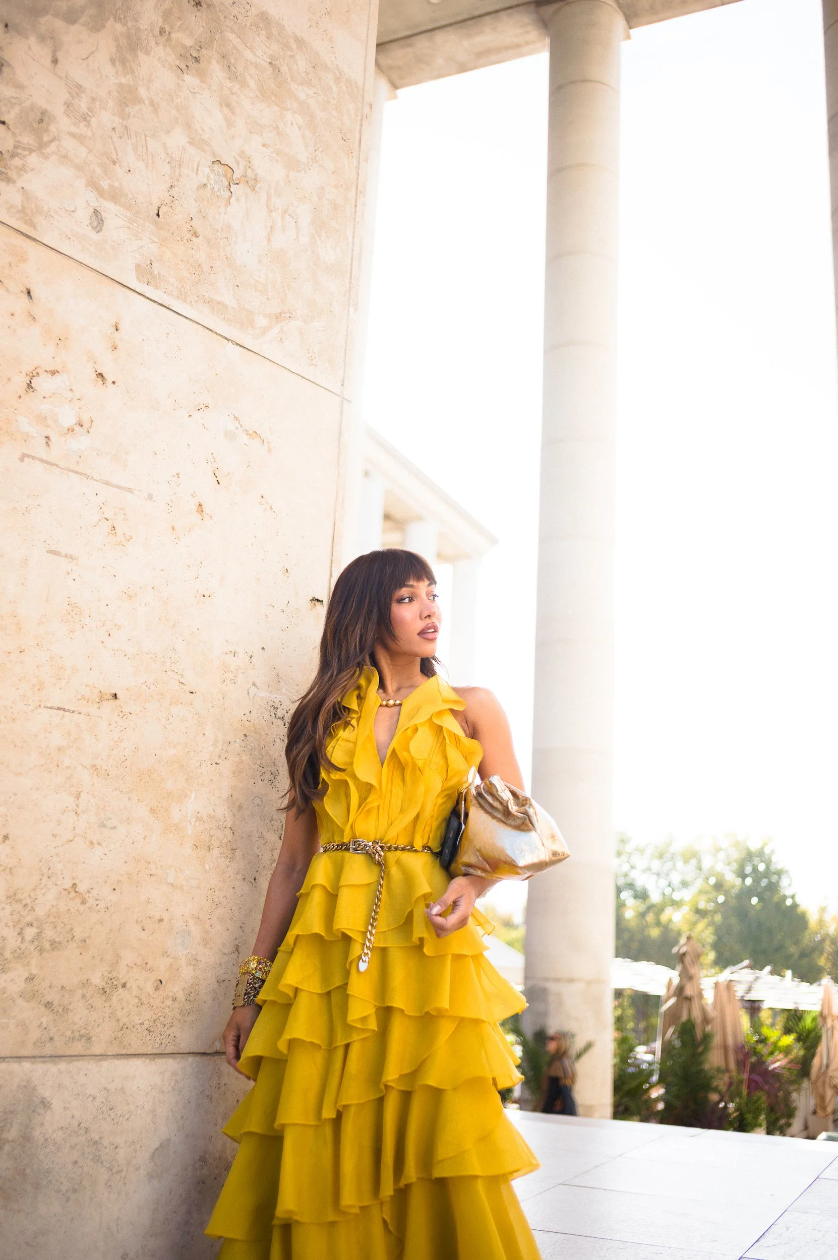 At Paris Fashion Week, A woman in a bright yellow ruffled dress stands outdoors, leaning against a stone wall, holding a large gold purse and wearing jewelry, with columns and trees in the background.