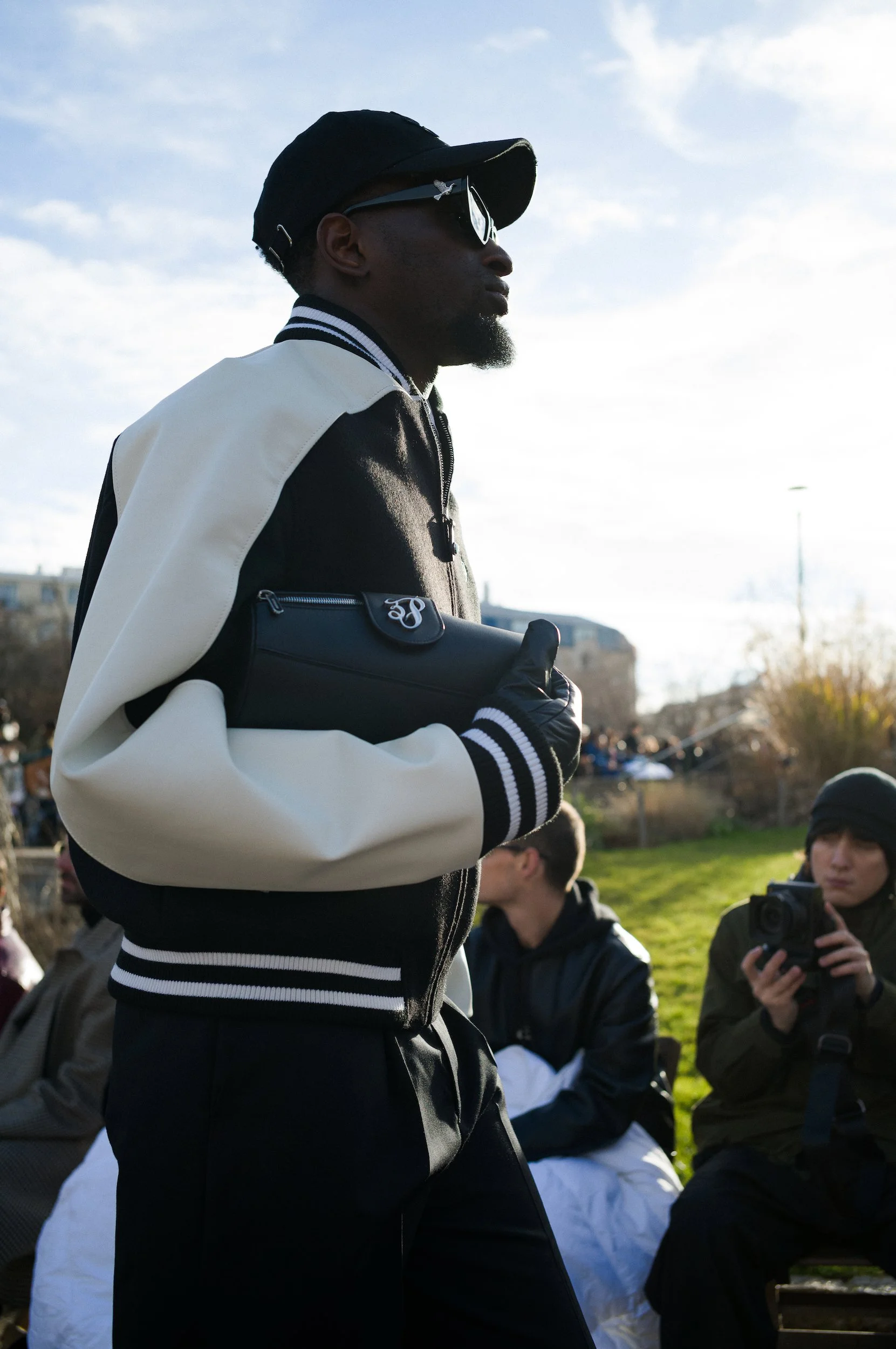 At Paris Fashion Week, A man wearing sunglasses, a black baseball cap, and a black and white varsity jacket stands outdoors with a group of people, some of whom are taking photos with cameras, on a grassy area under a partly cloudy sky.