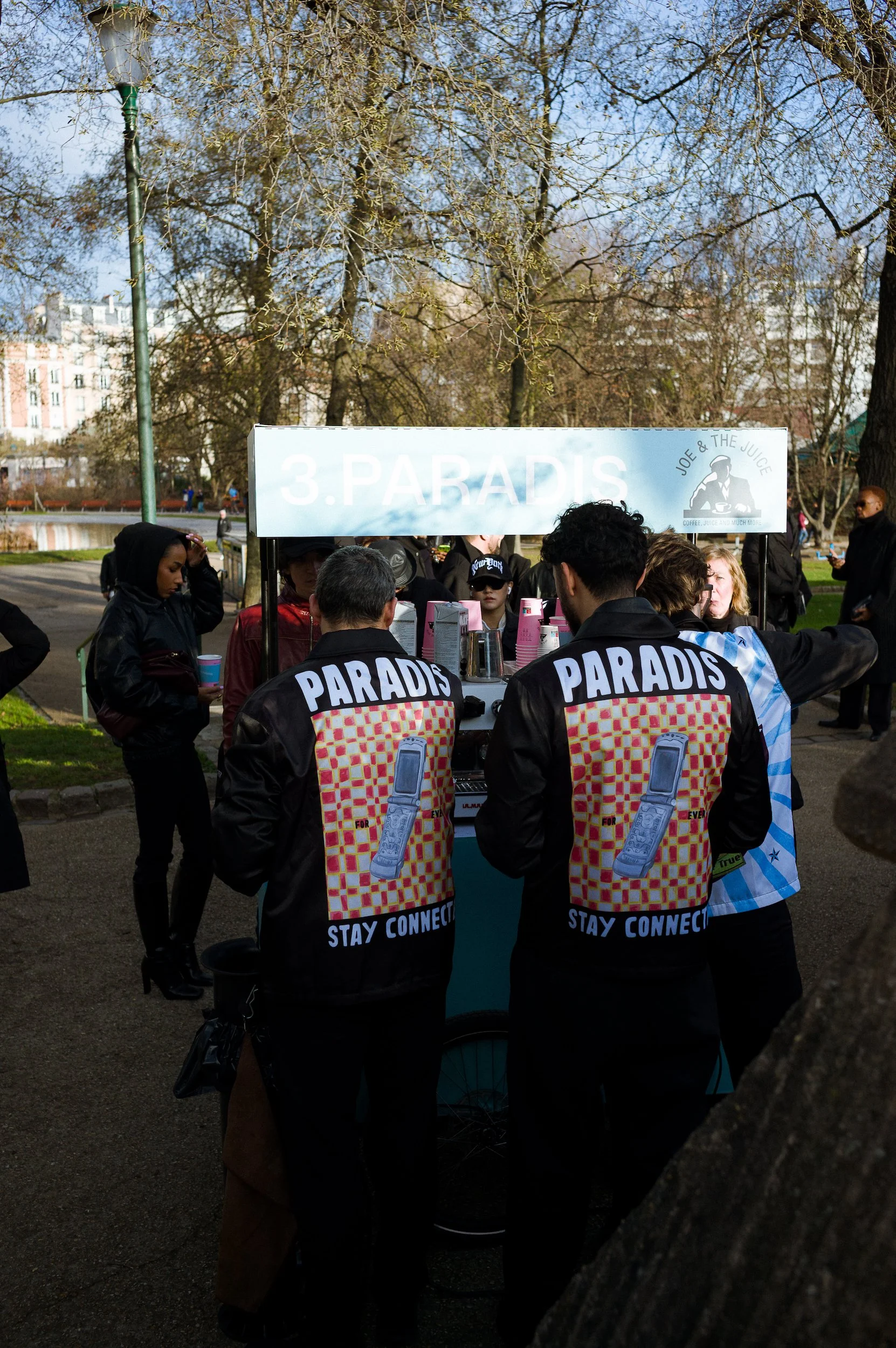 People gathered at a market stall labeled '3. PARADIS' with a logo reading 'Joe & The Juice'. The people are wearing jackets with 'PARADIS' print and the slogan 'STAY CONNECTED'. The setting appears to be a park with trees and buildings in the backgr