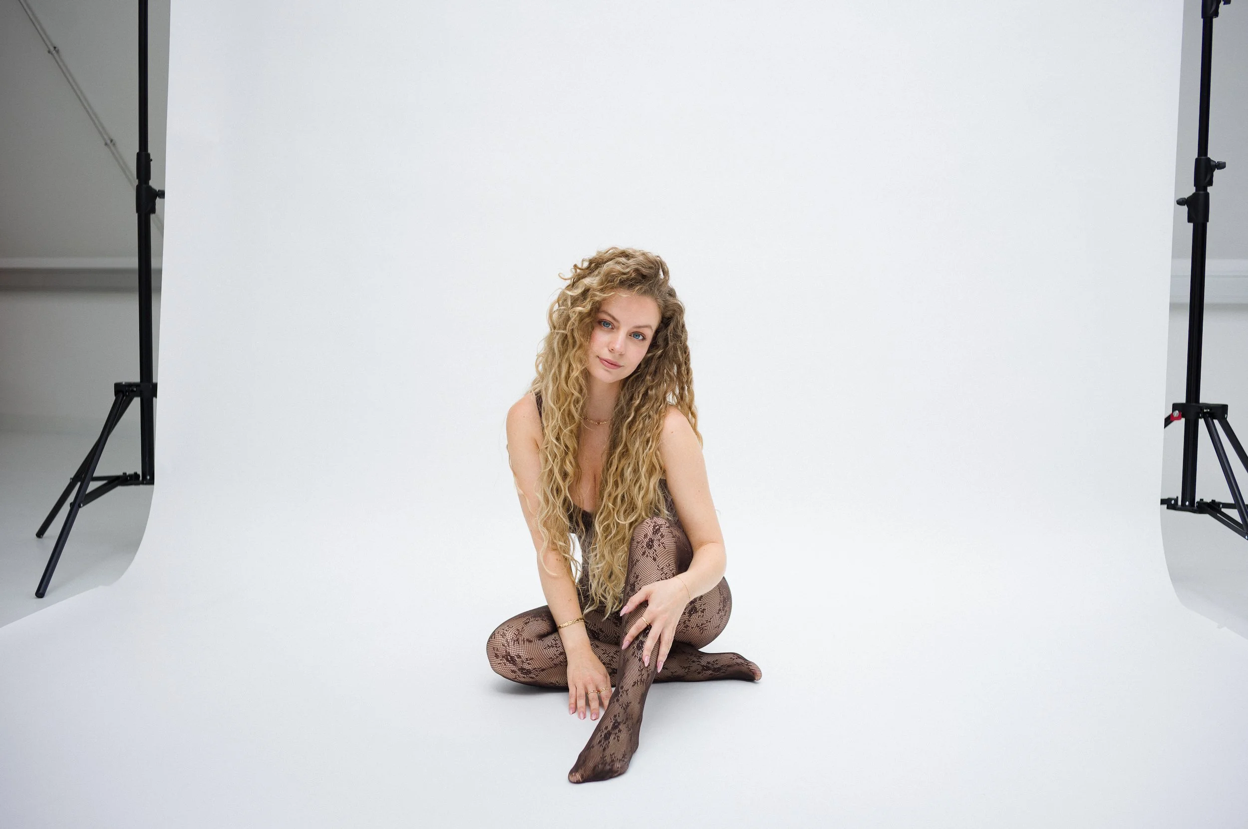 A woman with curly blonde hair sitting on a white photo studio floor in front of a white backdrop, surrounded by photography equipment.