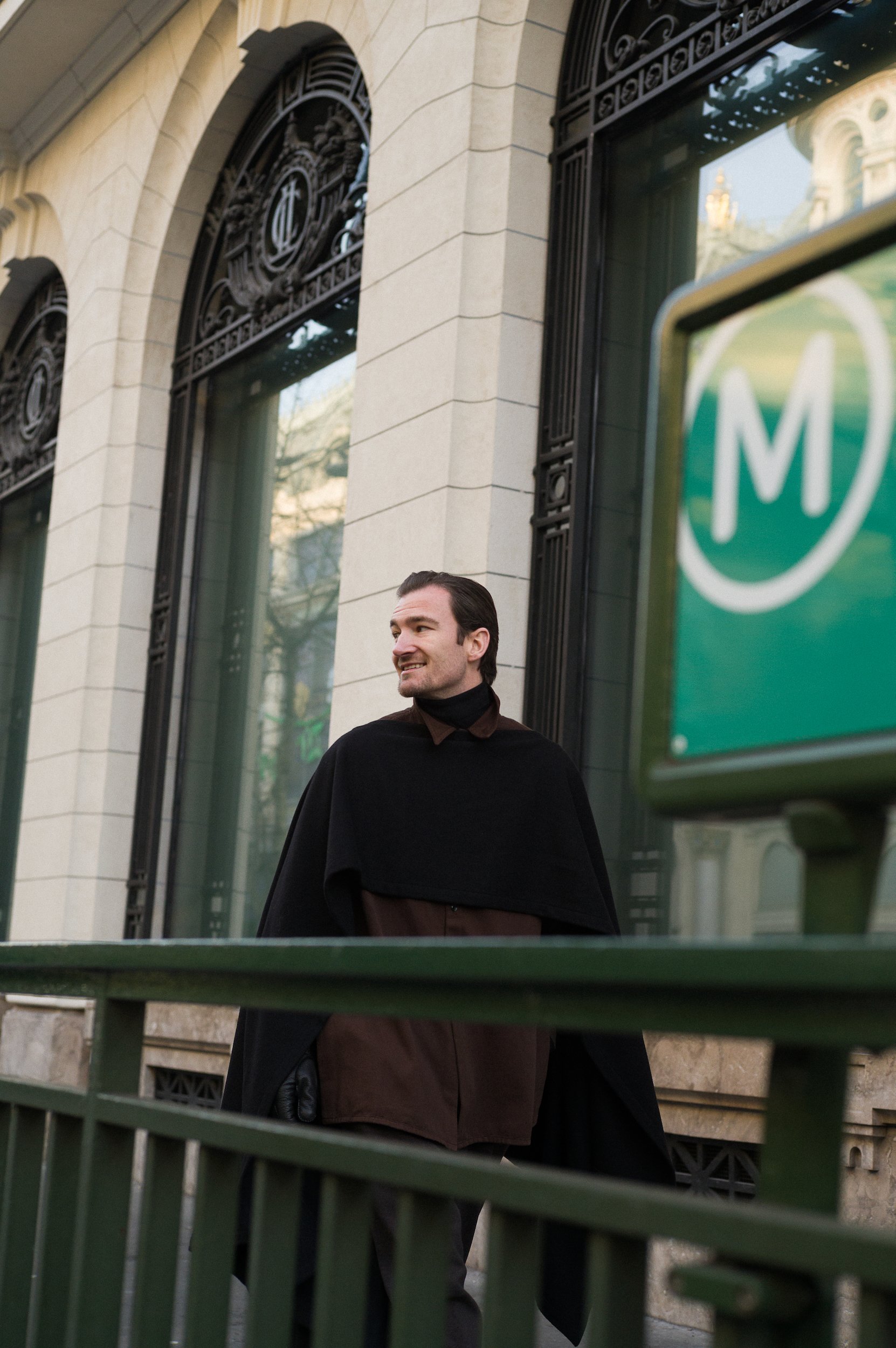 At Paris Fashion Week, A man with dark hair and a beard smiling, wearing a dark coat and brown pants, standing outside in front of a building with ornate black metal grates and large windows, with green metal railings in the foreground.