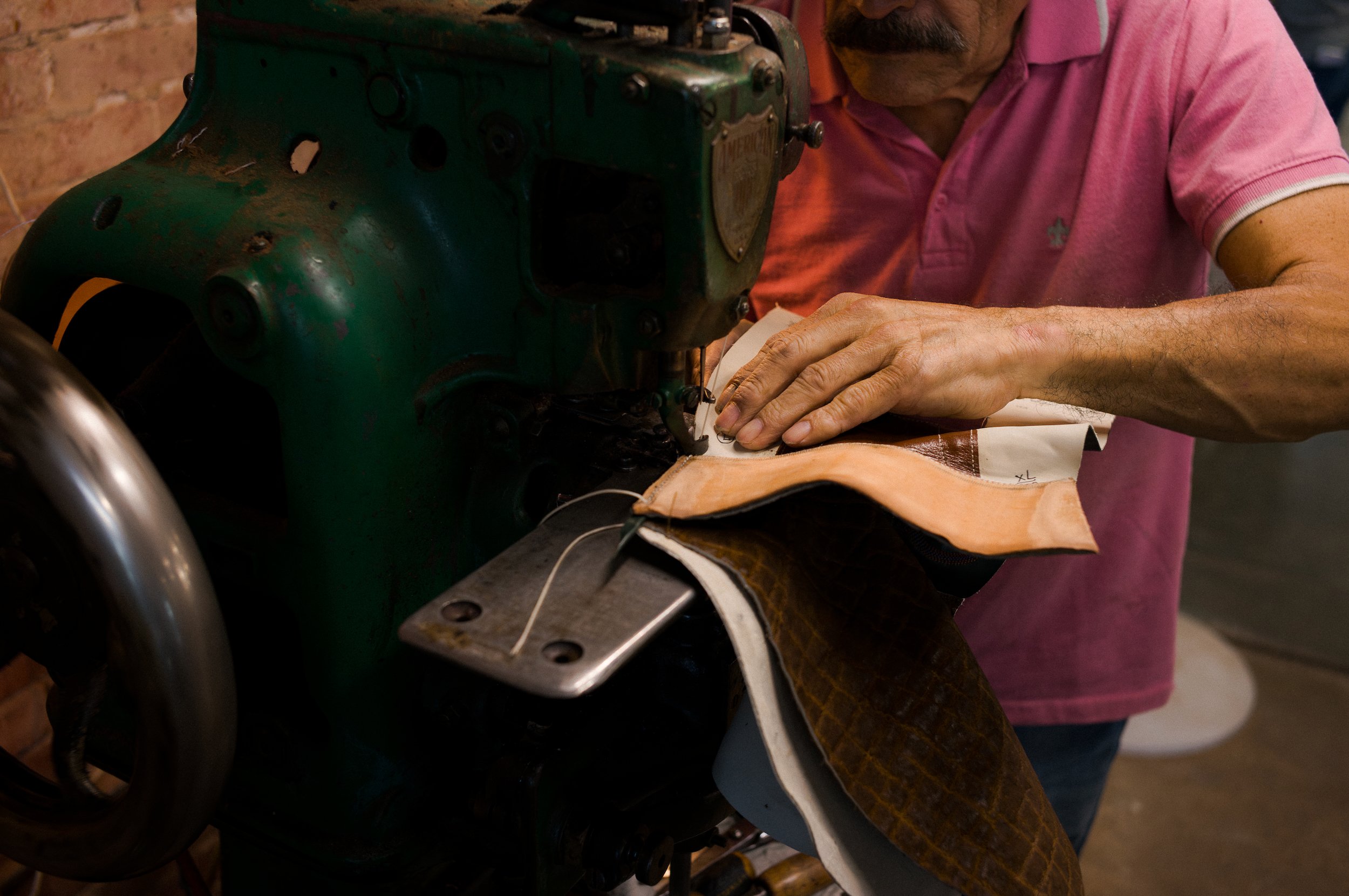 A person wearing a pink shirt working on a vintage green machine. The person's hands are on a piece of fabric, possibly sewing or repairing. The background has a brick wall.
