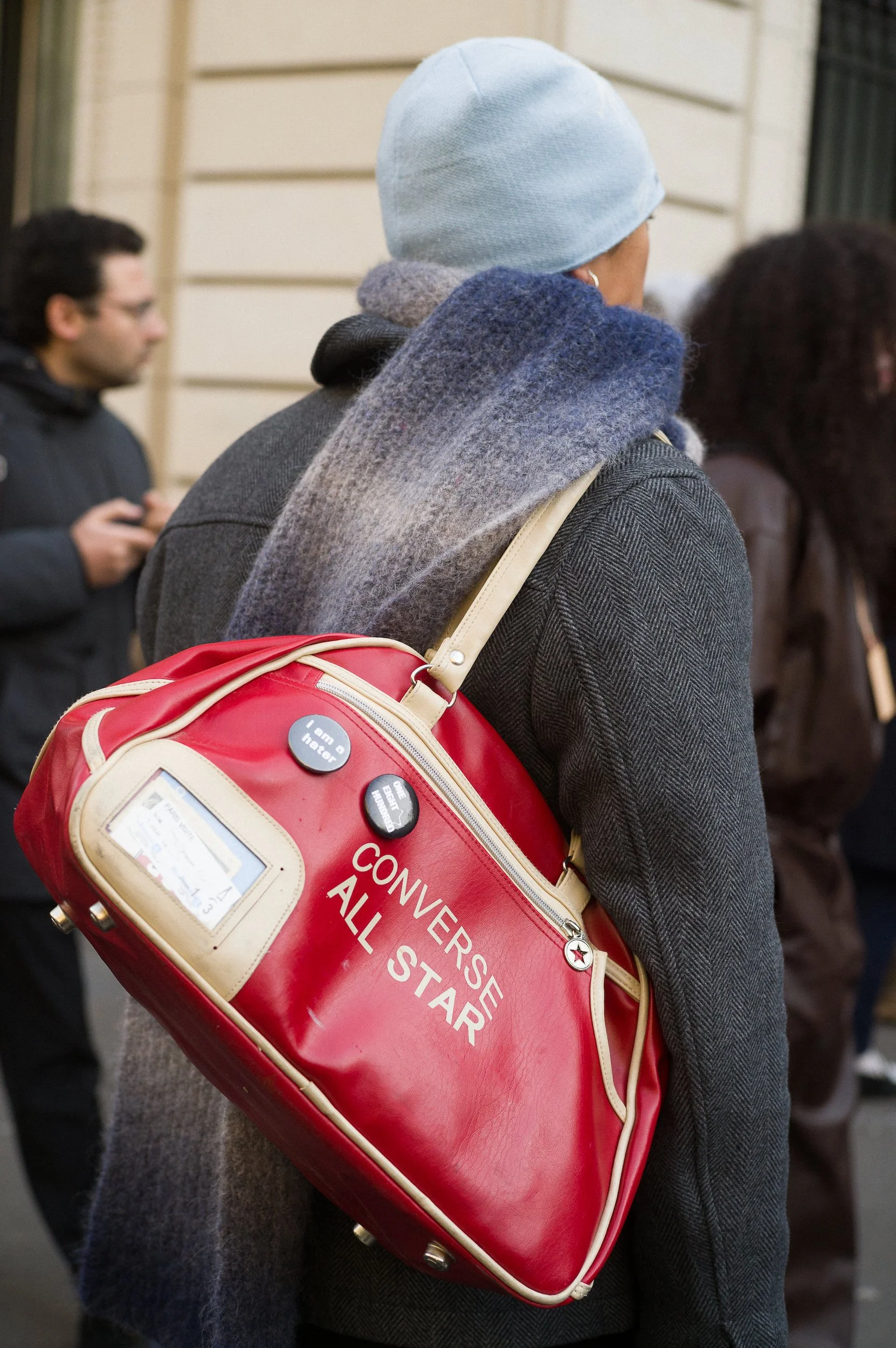 At Paris Fashion Week, Person wearing a light blue beanie, dark gray coat, and carrying a red Converse All Star gym bag with buttons and a small screen, standing among other people in an indoor setting.