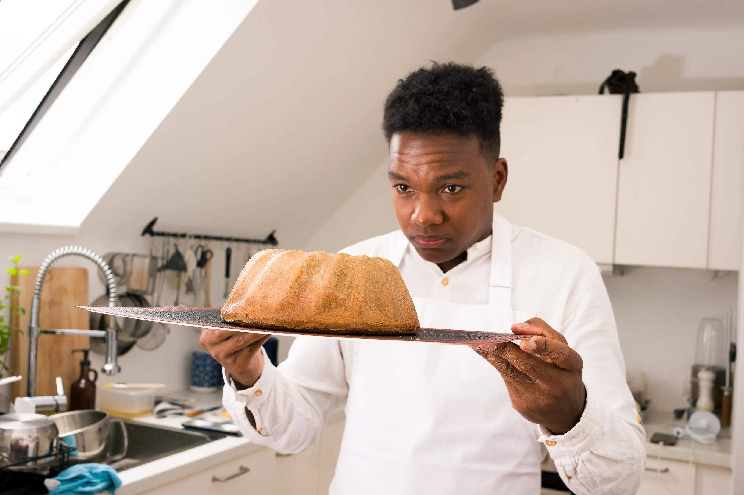 A chef in a white shirt holding a large baked loaf of bread on a silver tray in a kitchen.