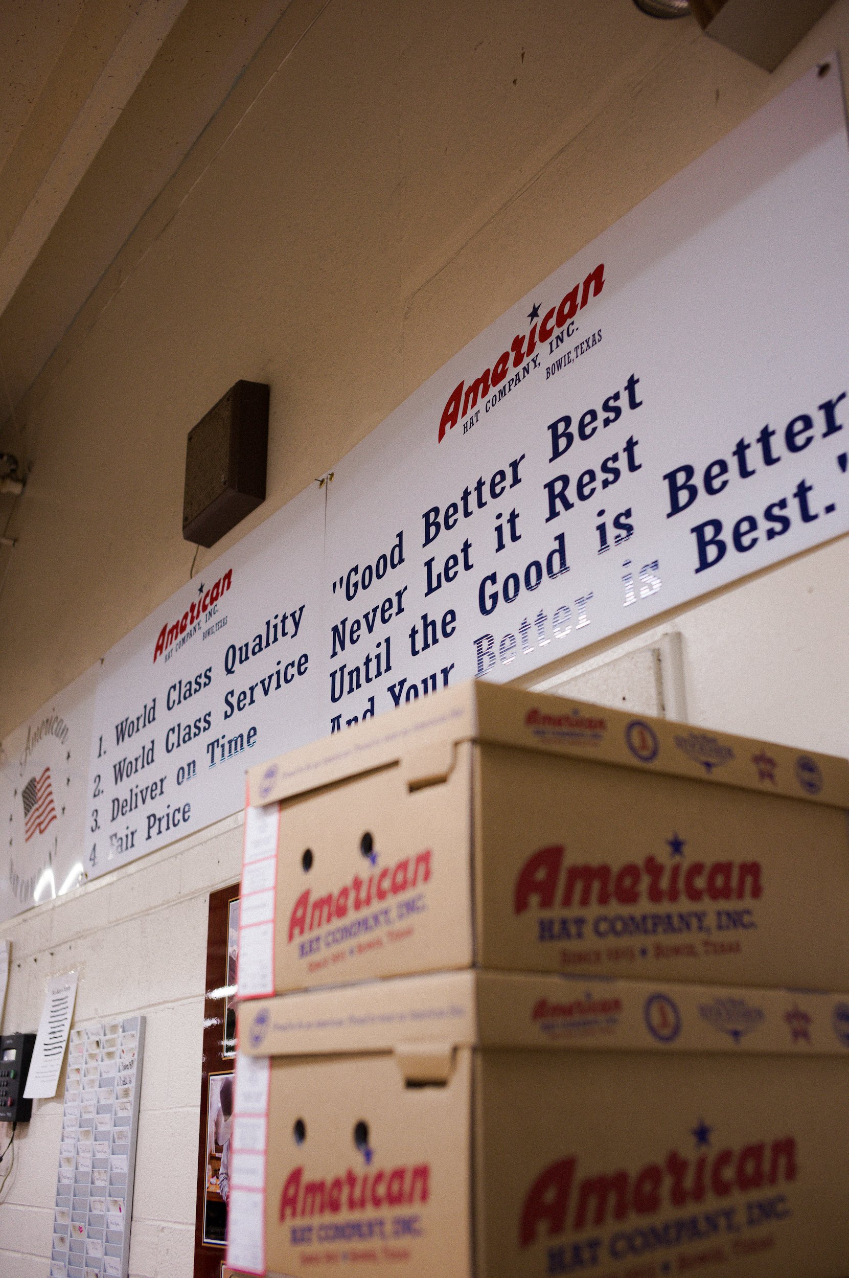 Close-up of a cardboard box labeled 'American Hat Company, Inc.' in front of a wall with handwritten notes and a bulletin board.