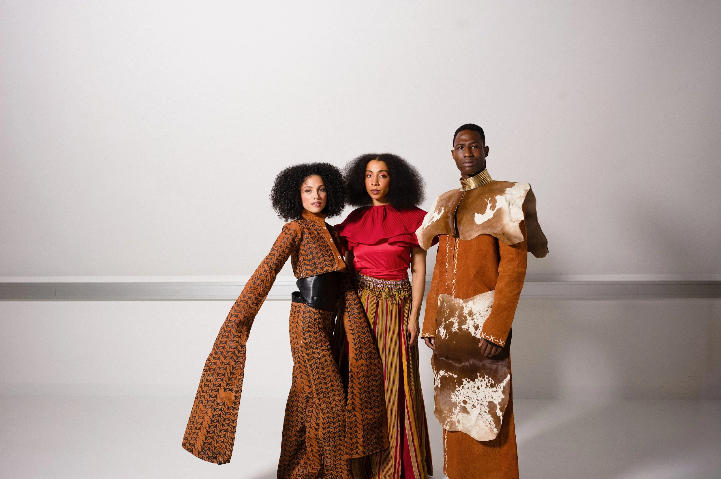 Three models standing against a plain white background, wearing colorful, patterned clothing with diverse hairstyles and accessories.