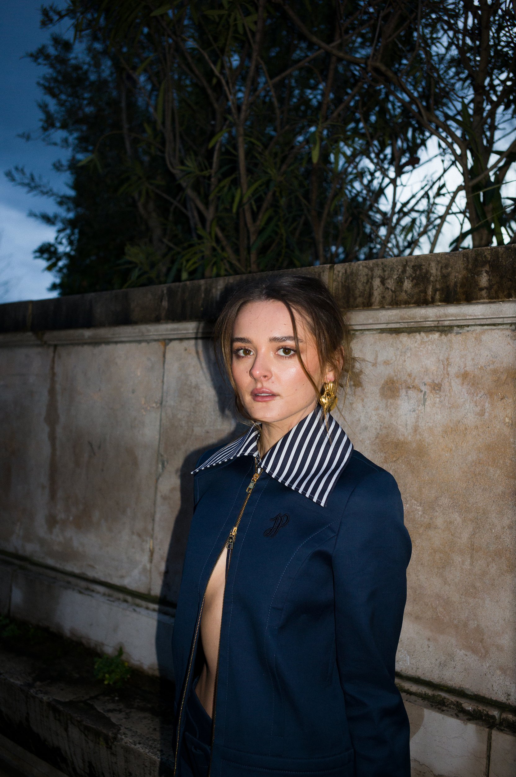 A young woman with brown hair, styled with loose waves, standing outdoors against a stone wall and greenery, wearing a navy blue jacket with a gold zipper and a striped shirt with a collar. The photo is taken during dusk or early evening.