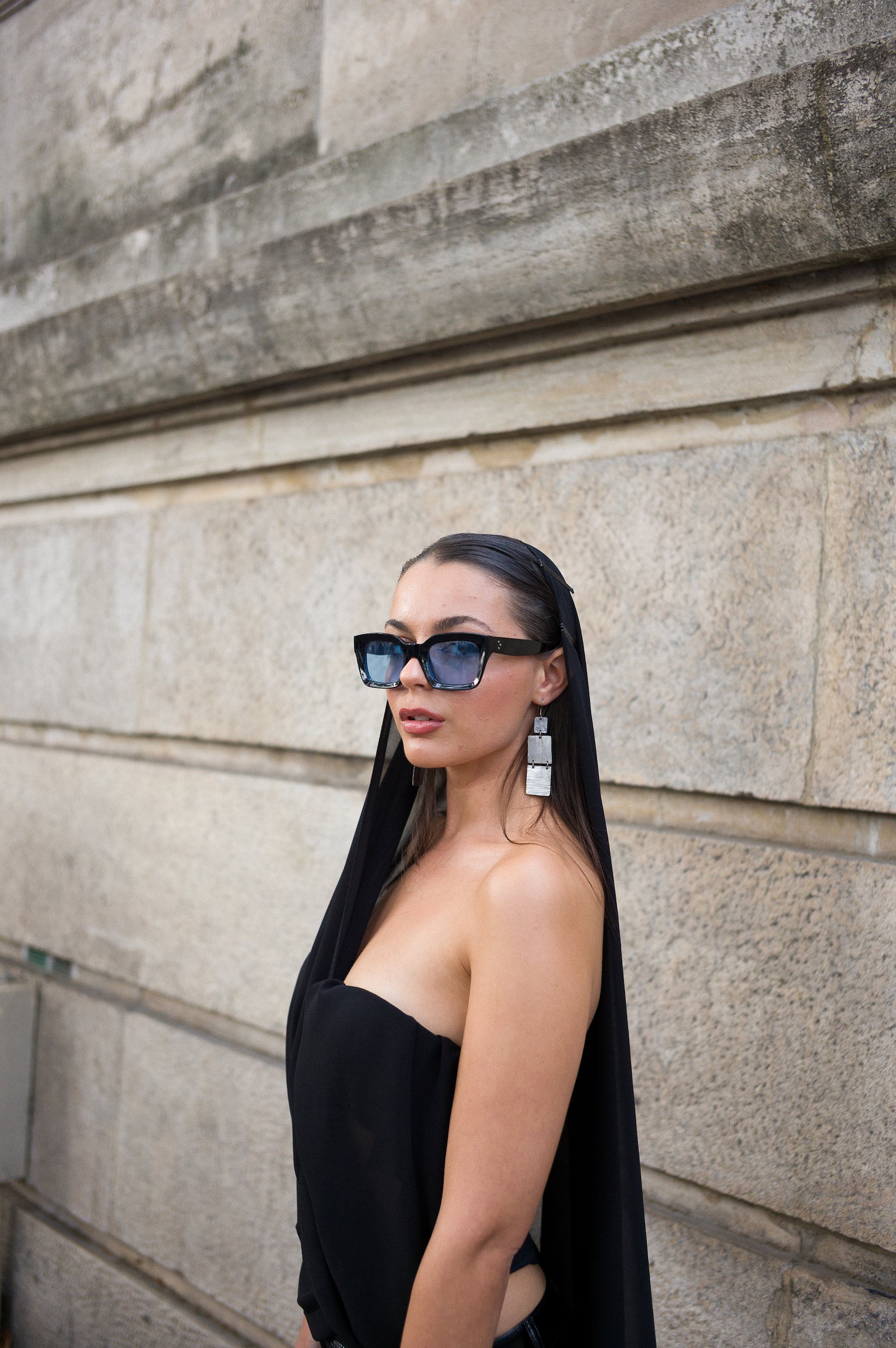 At Paris Fashion Week, A woman wearing large dark sunglasses with blue reflections, silver statement earrings, and a black off-the-shoulder outfit, standing against a textured stone wall.