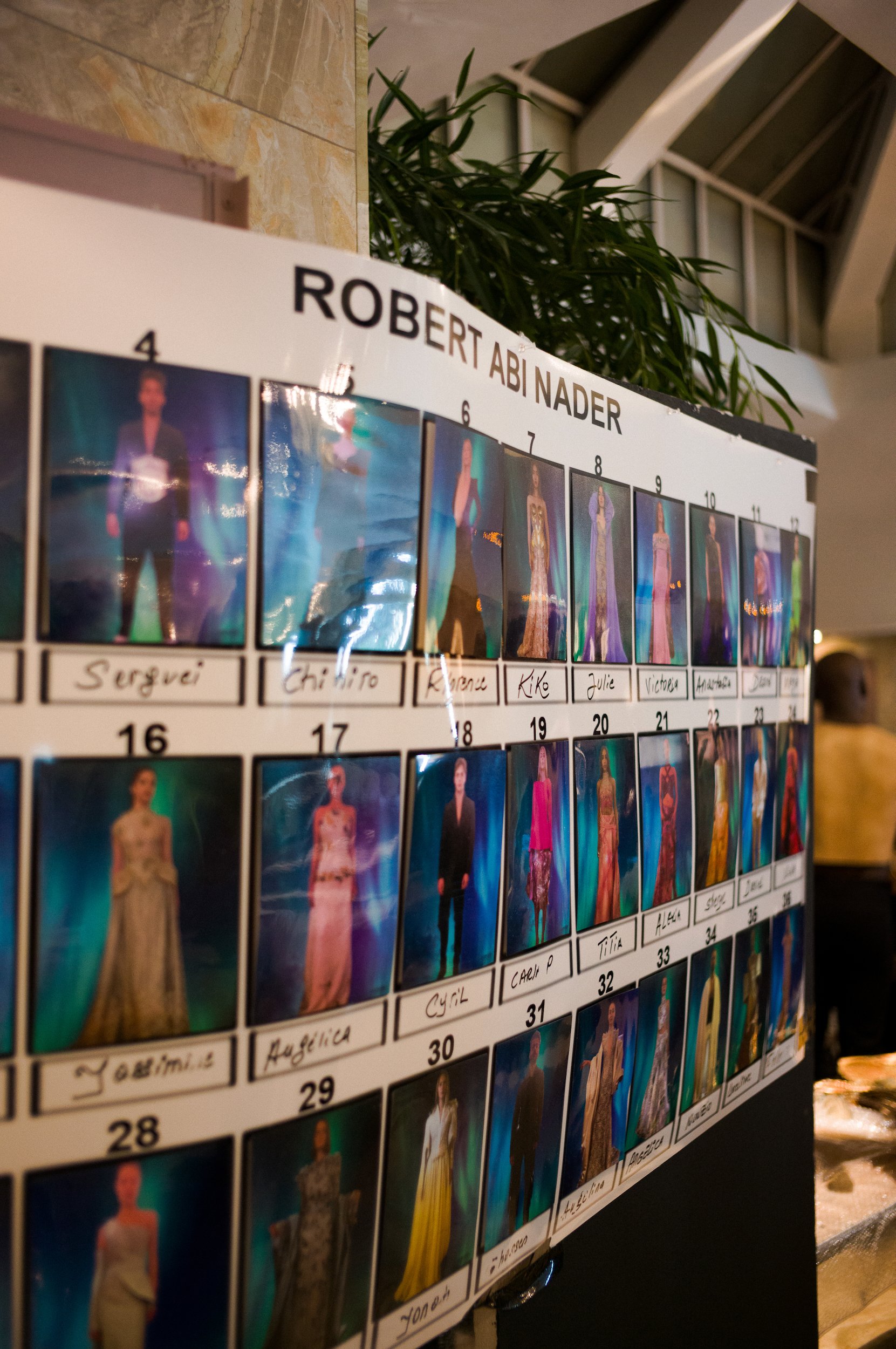At Paris Fashion Week, A photo of a display board with photographs of people dressed in formal attire, numbered and labeled with names, titled 'ROBERT ABI NADER'. The background includes a houseplant and part of a ceiling with beams.
