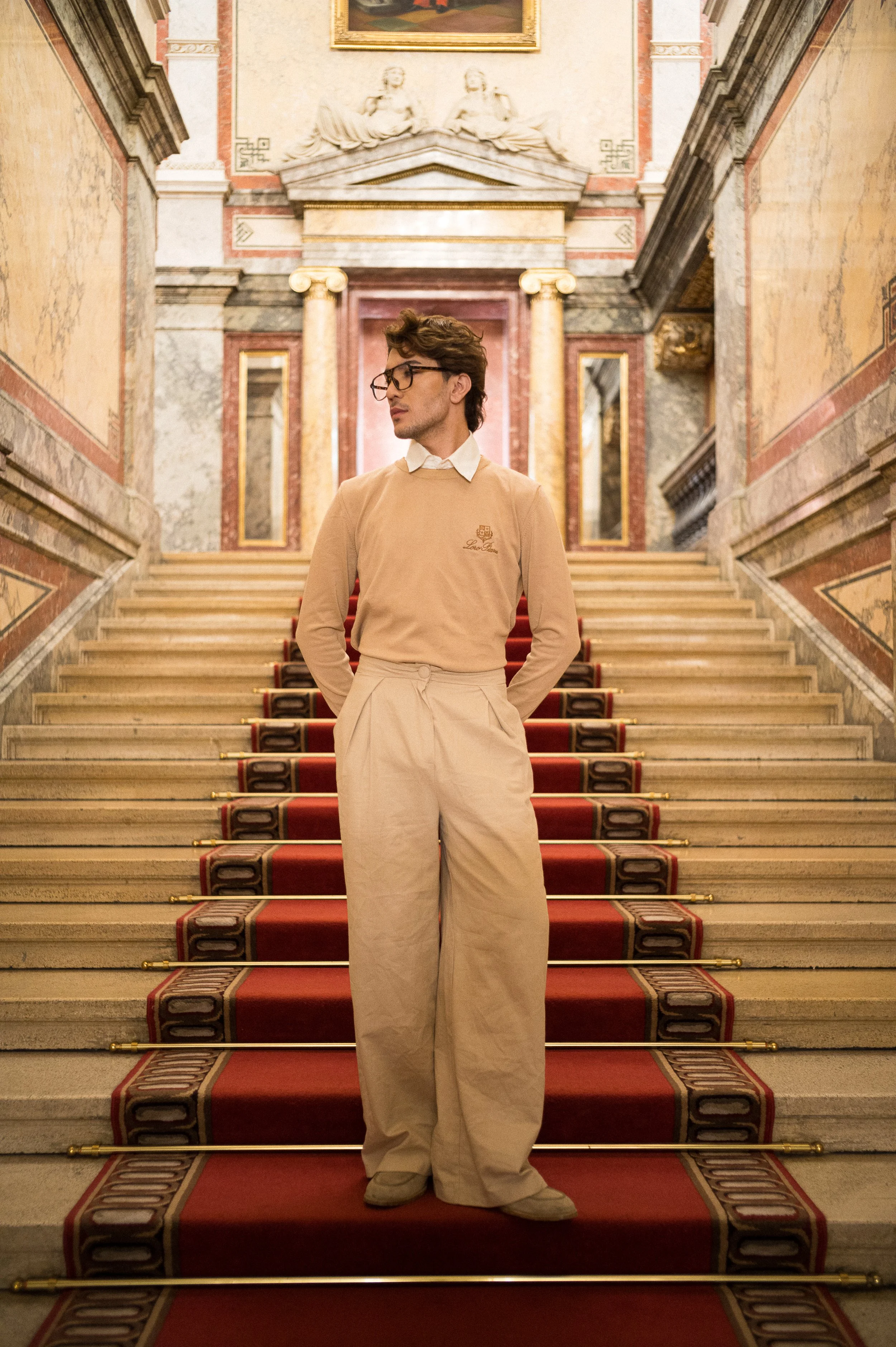 A young man with glasses, wearing a beige sweater and beige pants, standing on a grand staircase inside a historic building with ornate marble walls, pillars, and gold accents.