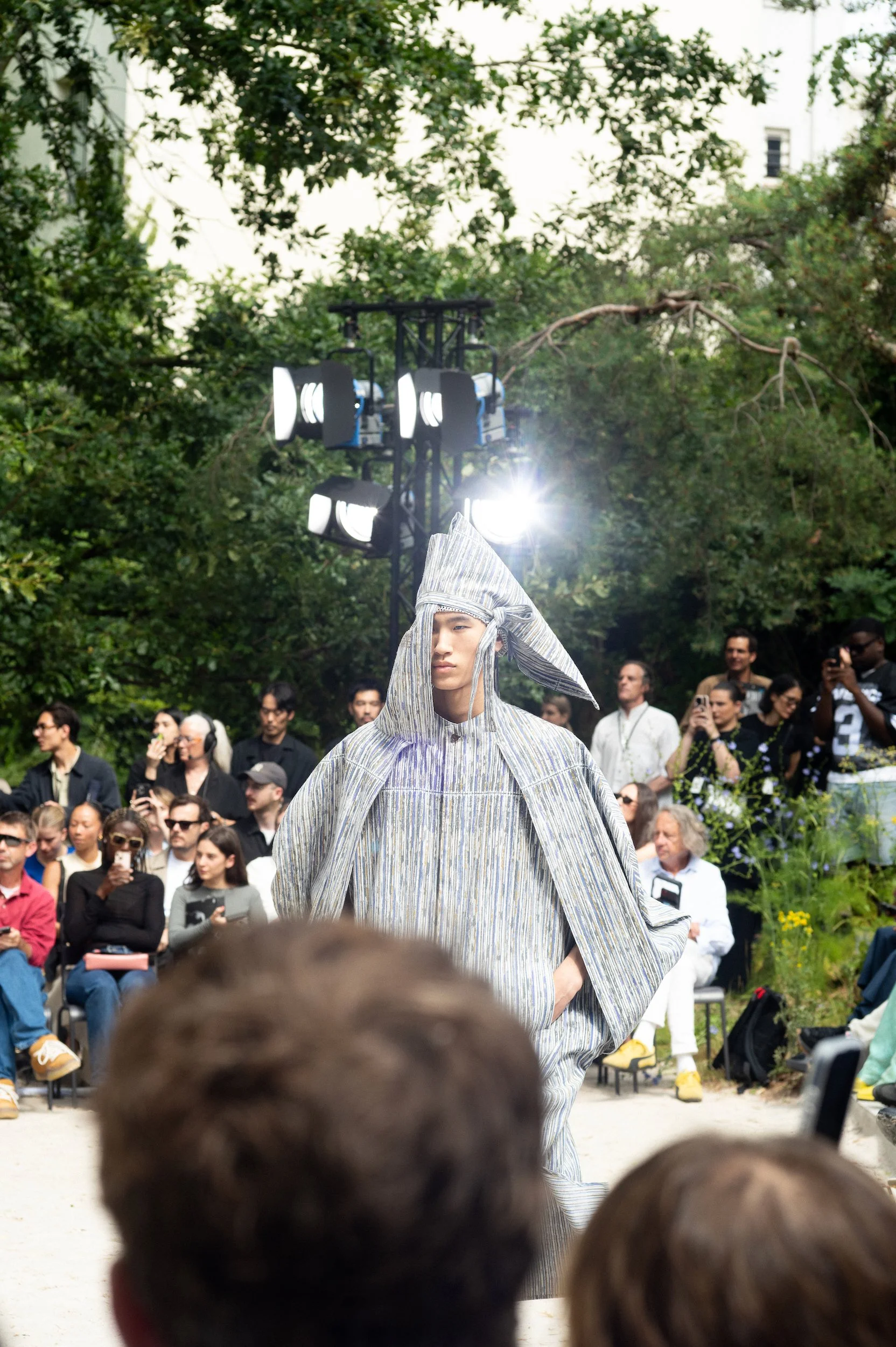 At Paris Fashion Week, A person walking the Issey Miyake runway wearing a gray, striped, loose-fitting outfit and a matching head covering, during a fashion show with an audience watching, and trees in the background.