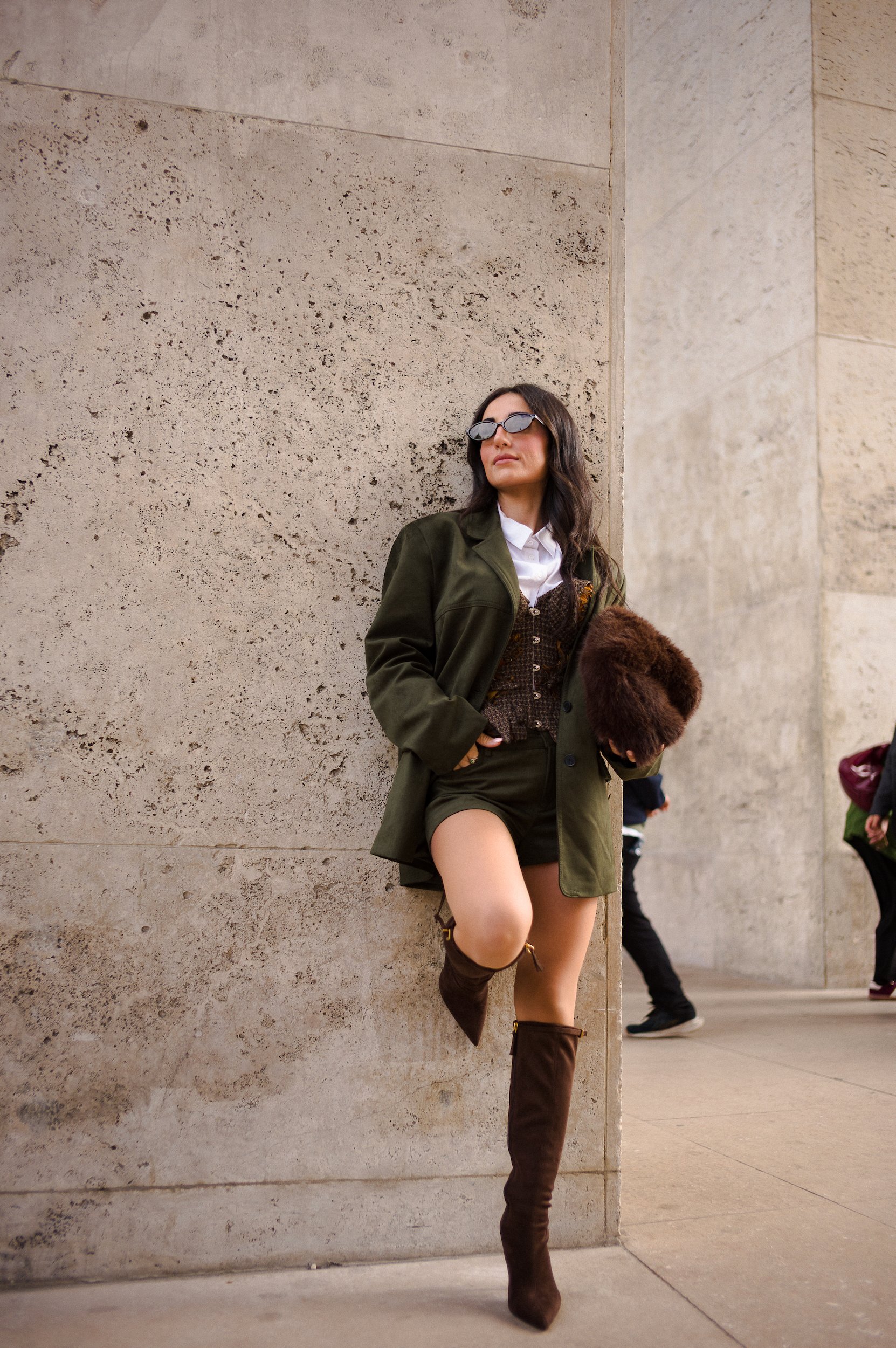 At Paris Fashion Week, A woman wearing sunglasses, a white shirt, an olive green jacket, and a brown dress stands against a beige stone wall, holding a fur coat and wearing brown knee-high boots.