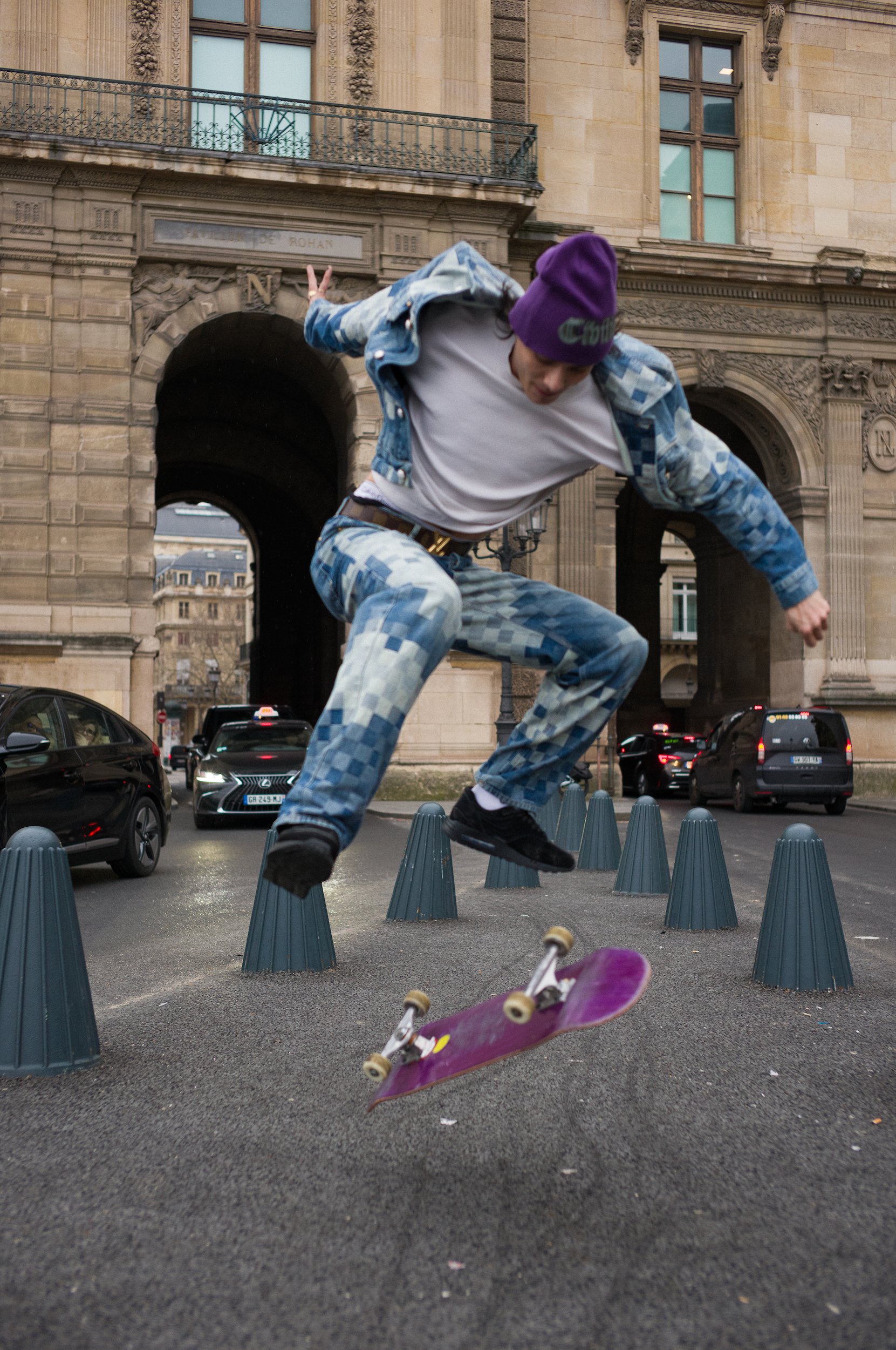 At Paris Fashion Week, A skateboarder wearing a purple beanie and blue patterned jacket is mid-air, jumping on a city street surrounded by traffic cones, with a historic building in the background.