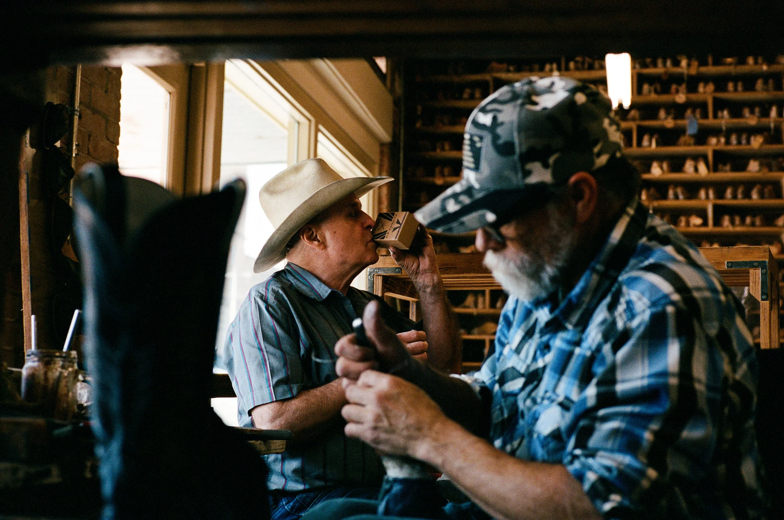 In ML Leddys, Two men, one wearing a cowboy hat and the other wearing a camouflage cap, are sitting at a workbench, one has a box of nails to his mouth in a rustic interior with shelves filled with boot patrons in the background.