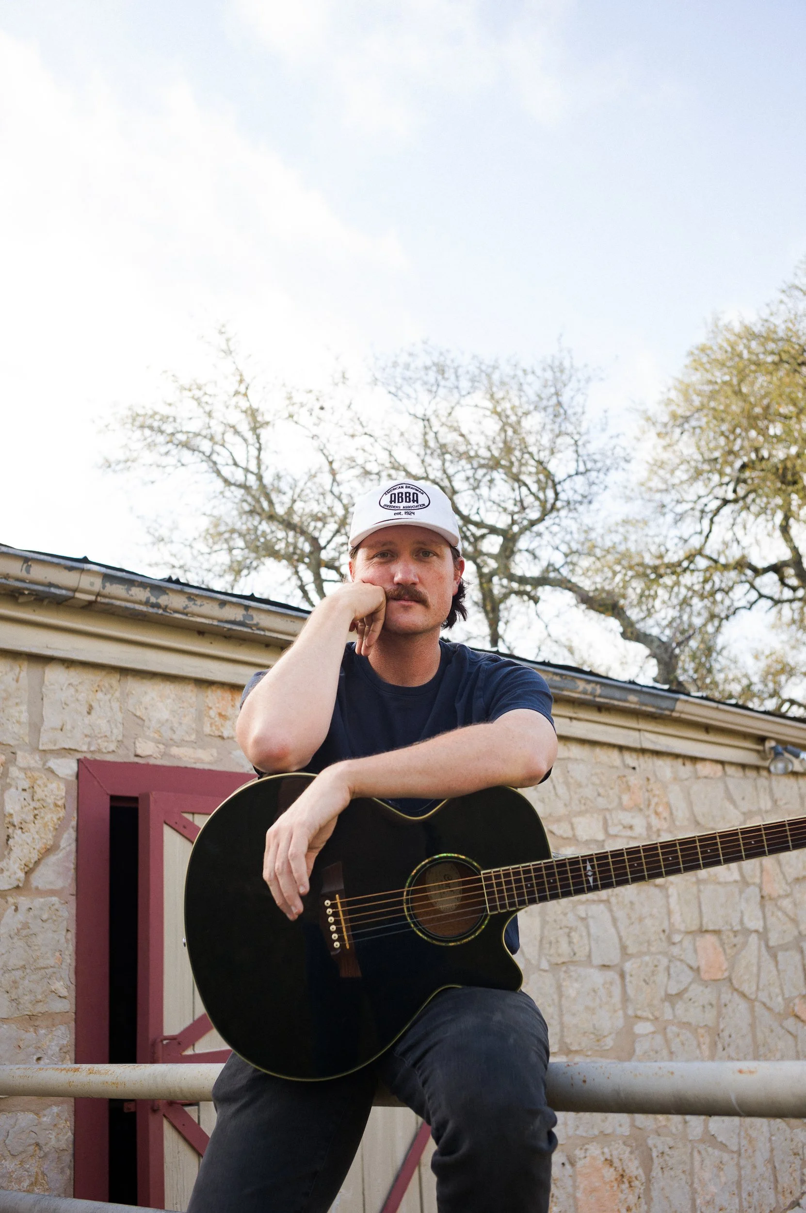 A man with a mustache wearing a white cap and a black shirt, sitting outside with a guitar, leaning on his left arm, looking at the camera, with trees and a brick building in the background.