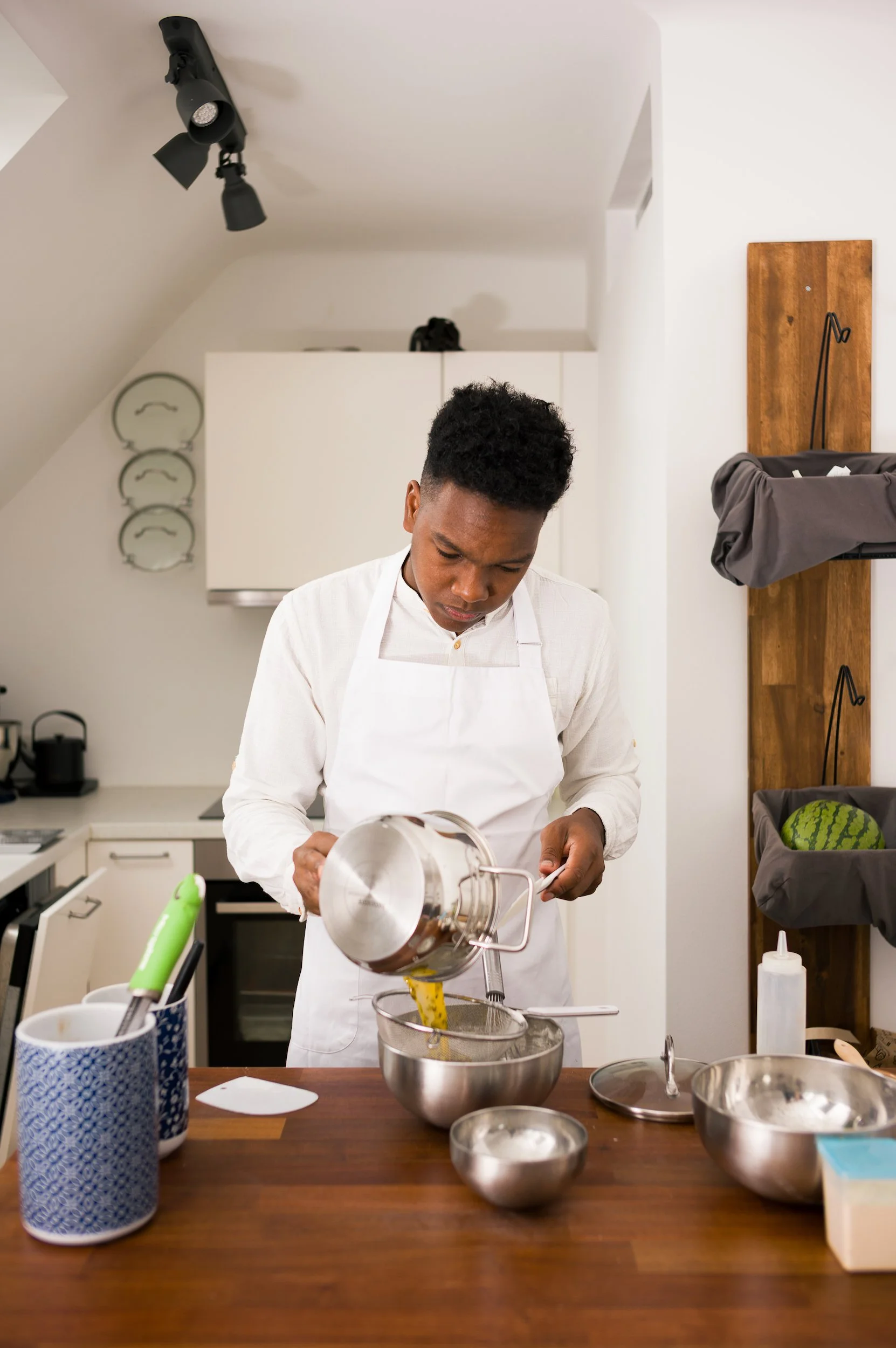 A young chef in a white apron is pouring batter into a mixing bowl in a modern kitchen.
