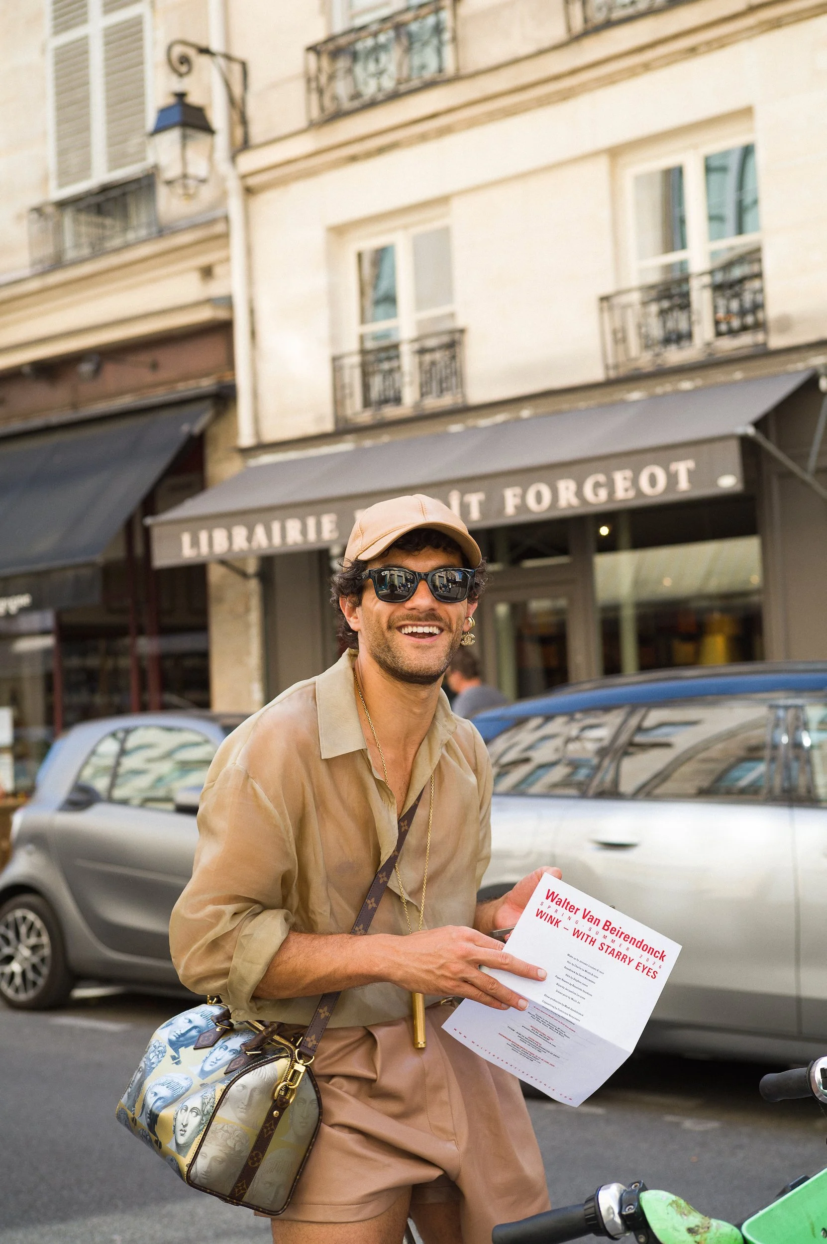 A smiling man wearing sunglasses, a beige cap, and a beige shirt holds a paper flyer on a city street. He has a patterned bag hanging from his shoulder. In Paris
