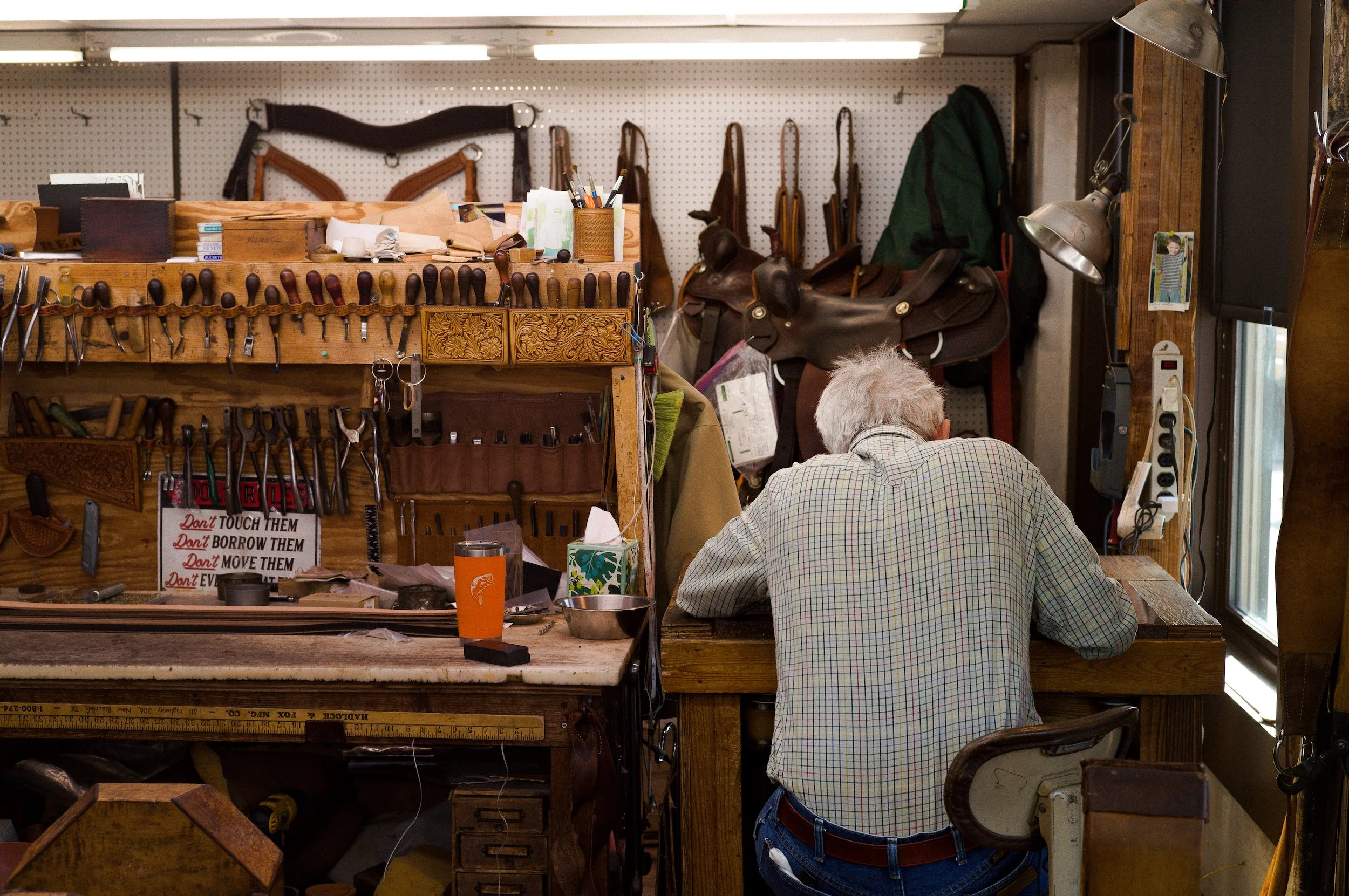 A workshop with various leatherworking tools, belts, and saddles on display. An elderly man with gray hair, wearing a checkered shirt and jeans, is seated at a workbench, working on a project. The workspace is cluttered with supplies, and there is a 
