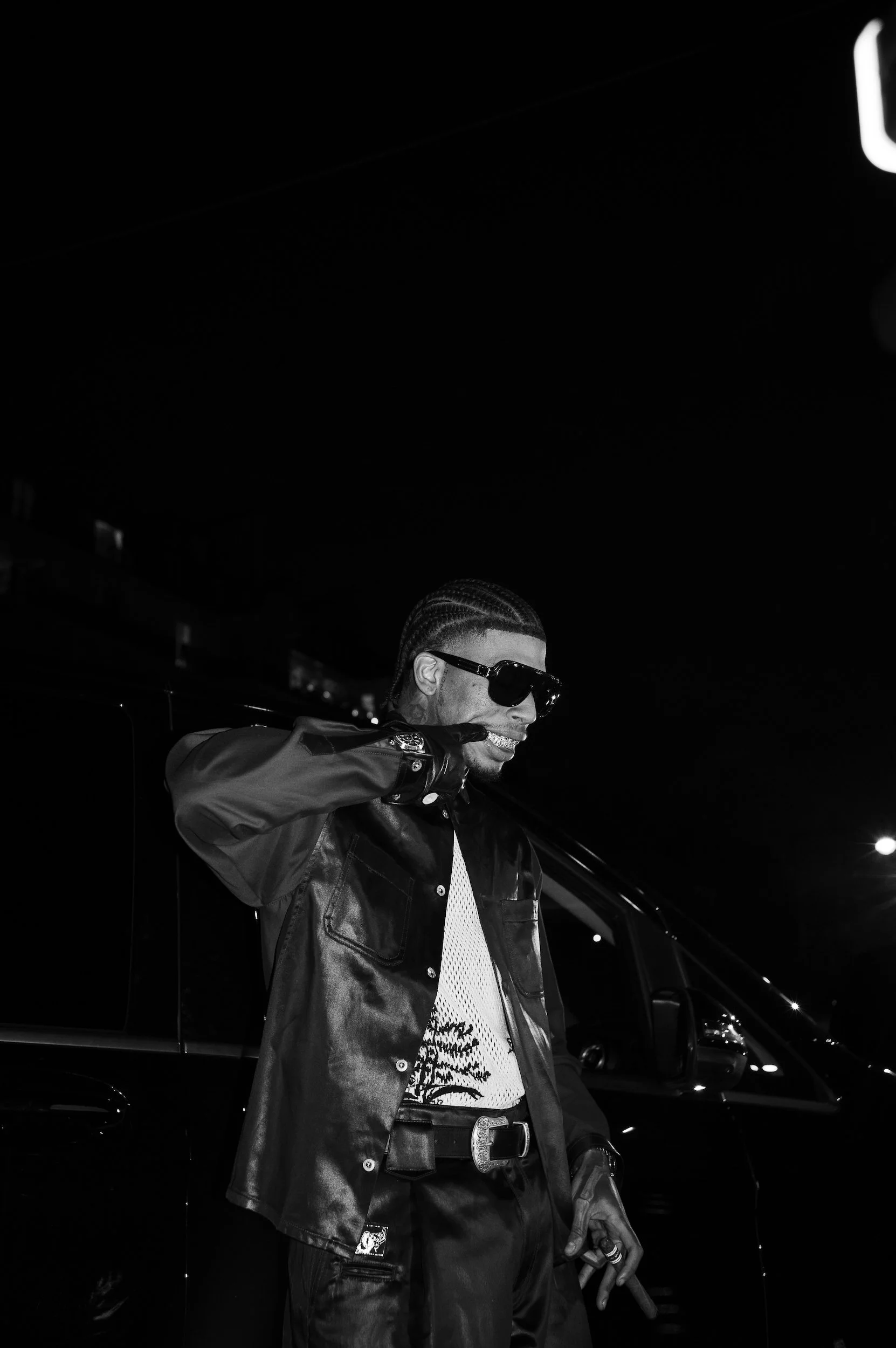 At Paris Fashion Week, A man with braided hair wearing sunglasses, a leather jacket, and a white patterned shirt standing next to a car at night, with a dark background and faint city lights.