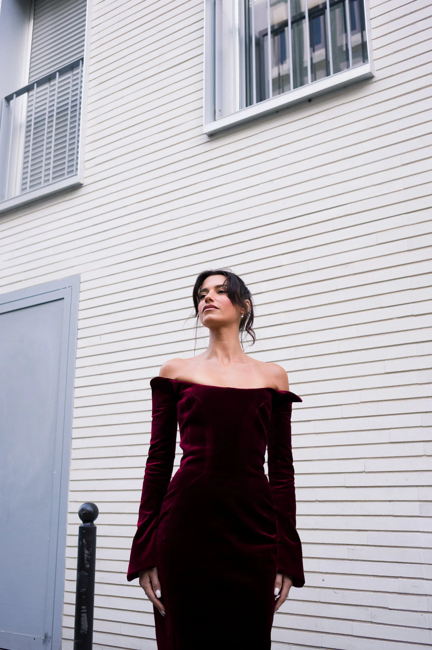At Paris Fashion Week, Woman in a strapless, long-sleeved, dark red velvet dress standing outside near a building with white siding and two windows.