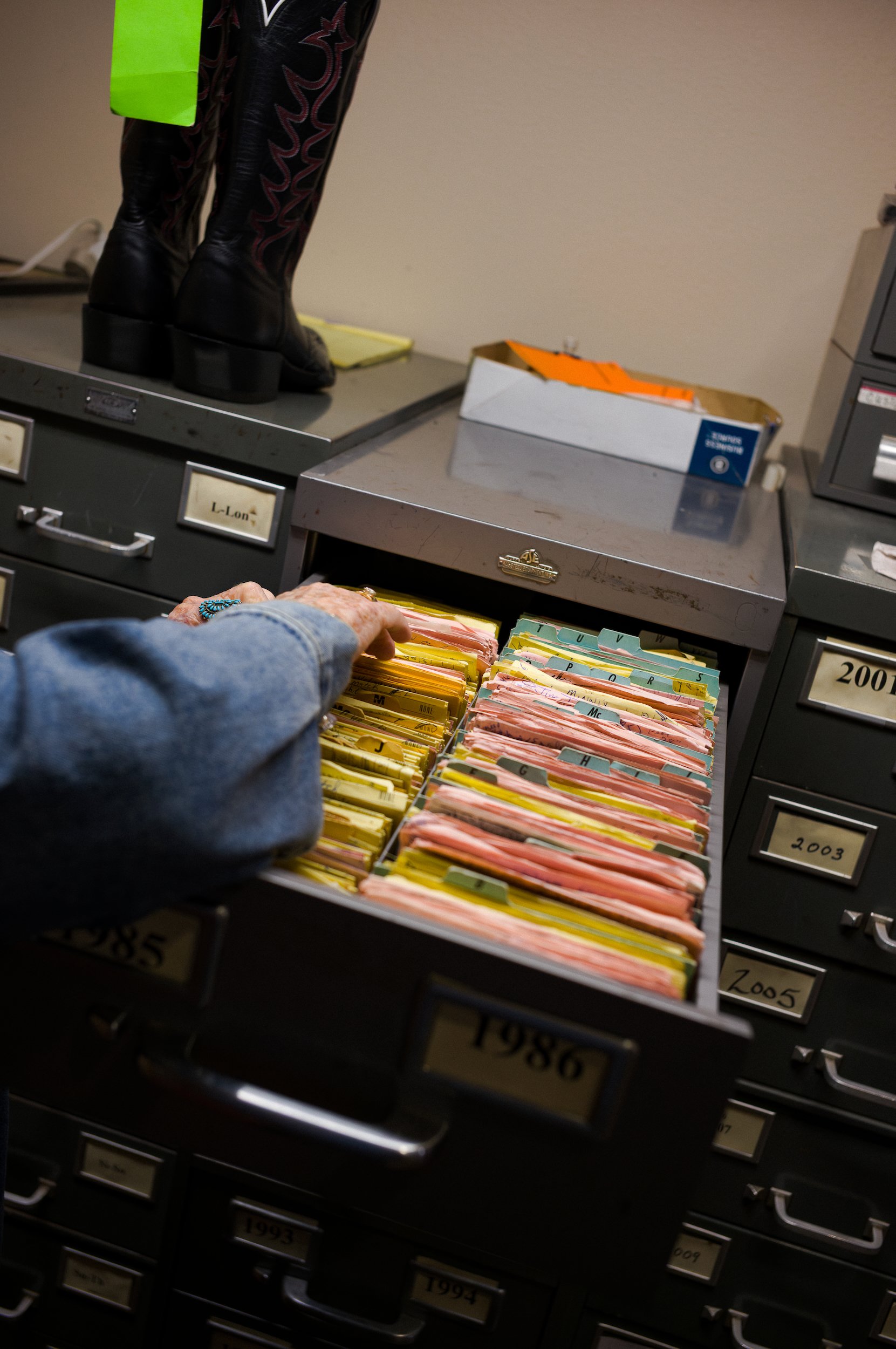 Close-up of a filing cabinet drawer filled with tabbed folders, some labeled with years, and a person's hand reaching for a folder. A pair of black cowboy boots is visible in the background.